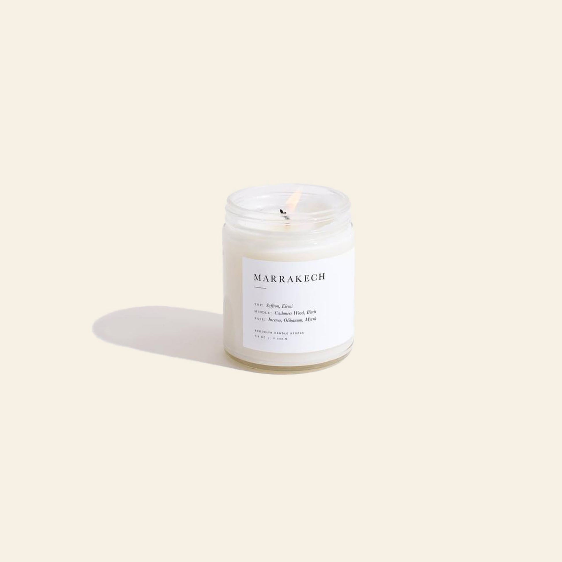 A lit Brooklyn Candle Studio Marrakech Jar Candle rests on a light beige surface, its glass jar casting a soft shadow. The white label features sleek black text listing notes like myrrh and saffron, plus ingredients.