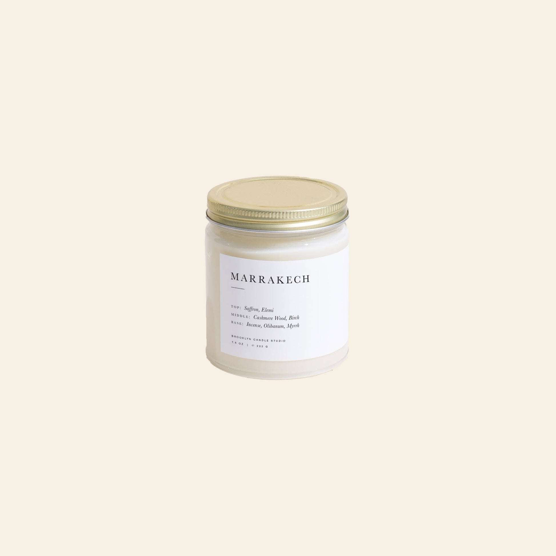 The Brooklyn Candle Studio Marrakech Jar Candle features a white glass jar with a gold lid and a minimalist label, infused with saffron and myrrh notes. It rests on a simple, light beige background.