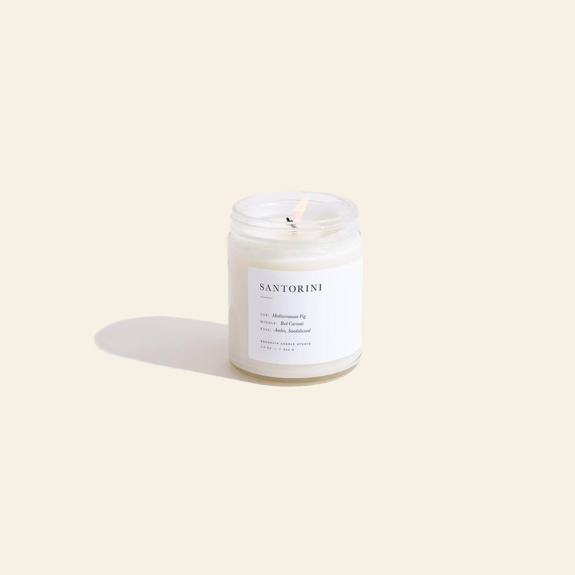 A Brooklyn Candle Studio Santorini Jar Candle glows in a clear glass jar on a beige surface, its wick radiating Mediterranean Fig, red currant, and amber to evoke the signature Greek Isles fragrance.