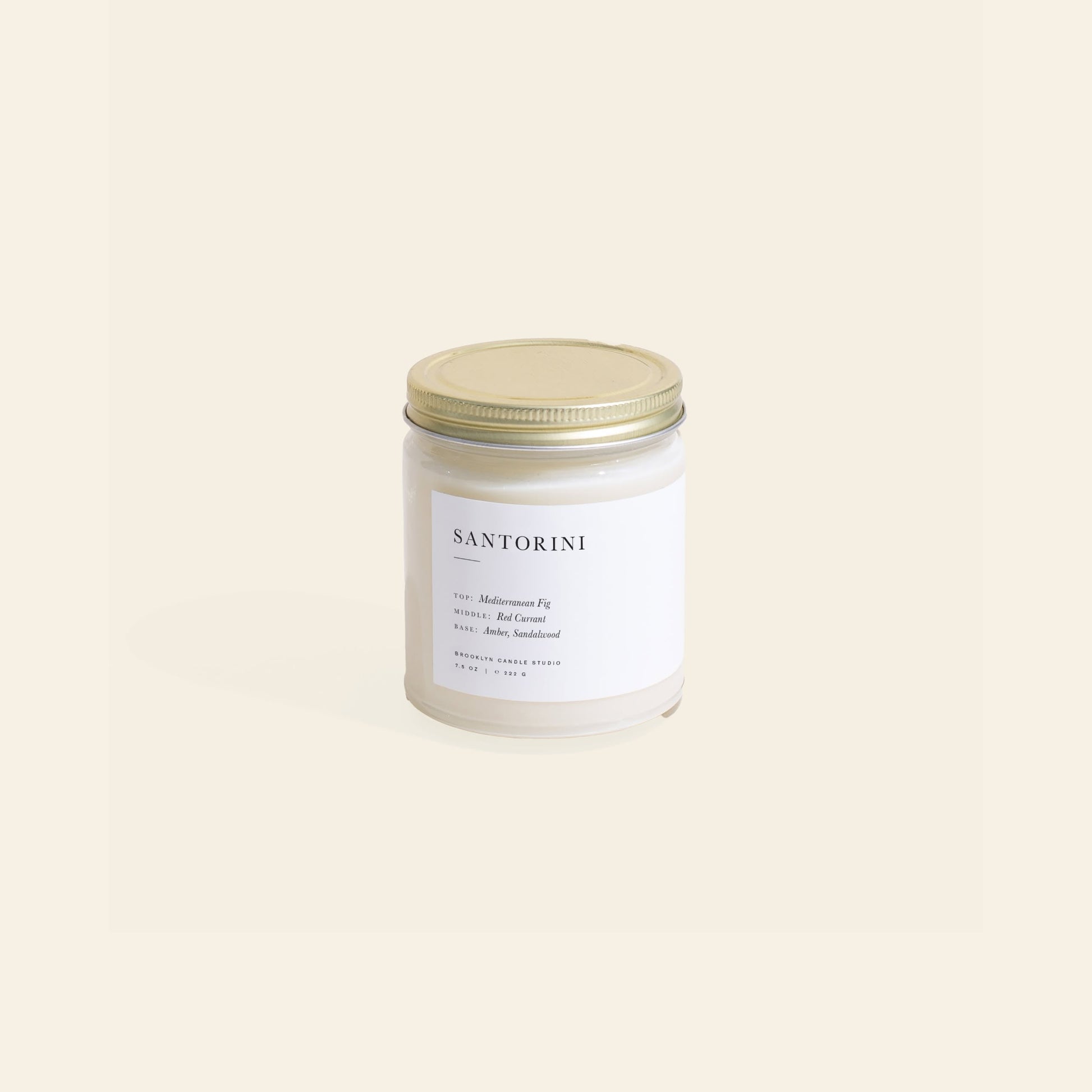 The Brooklyn Candle Studio Santorini Jar Candle features Mediterranean fig, red currant, amber, and sandalwood in a glass jar with a gold lid and white label—a Greek Isles-inspired scent set on a plain beige background.