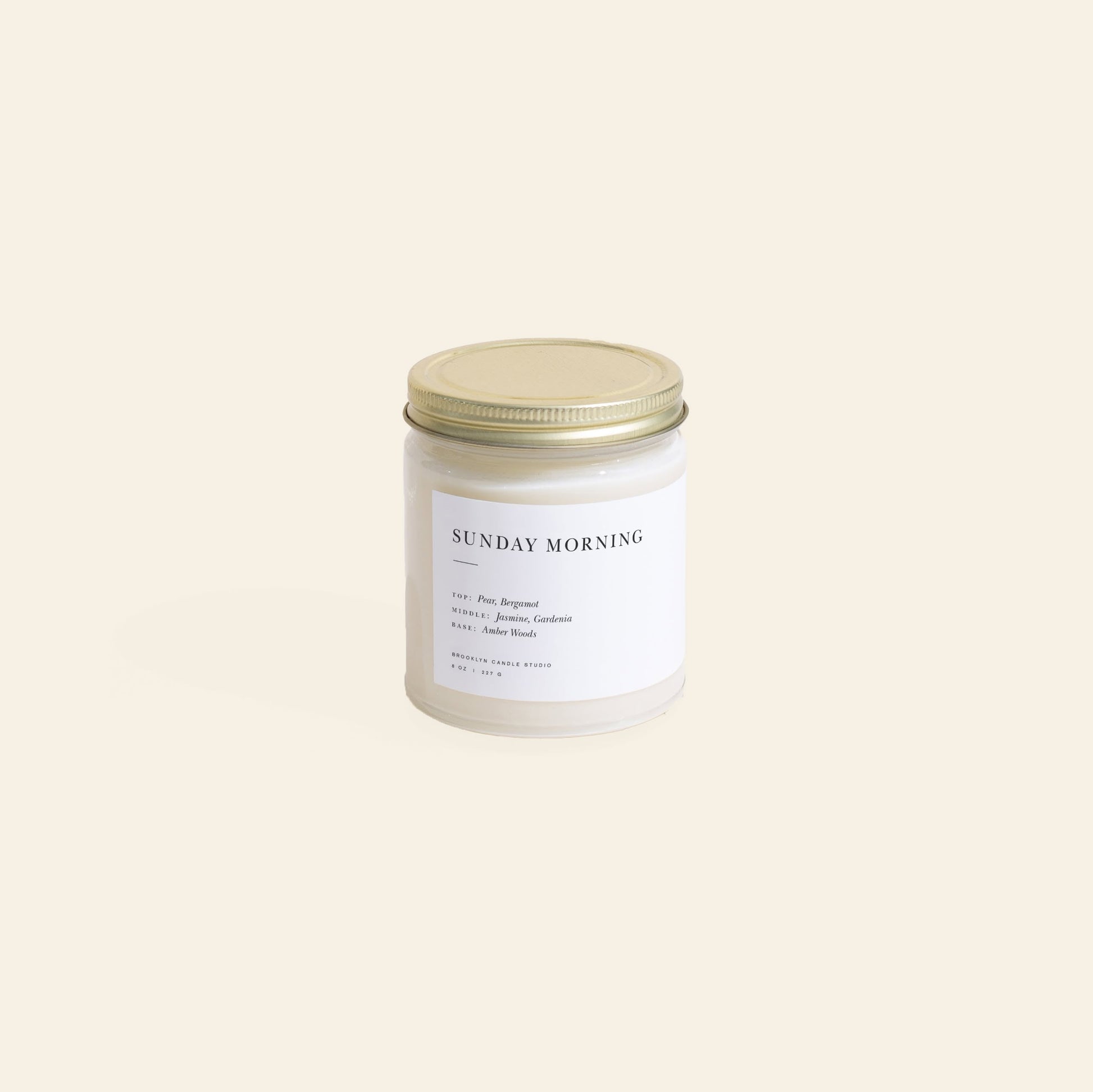 The Brooklyn Candle Studio Sunday Morning Jar Candle features a gold lid and label with notes of pear, bergamot, jasmine, gardenia, and amber woods. The glass jar candle is set against a simple light cream background.