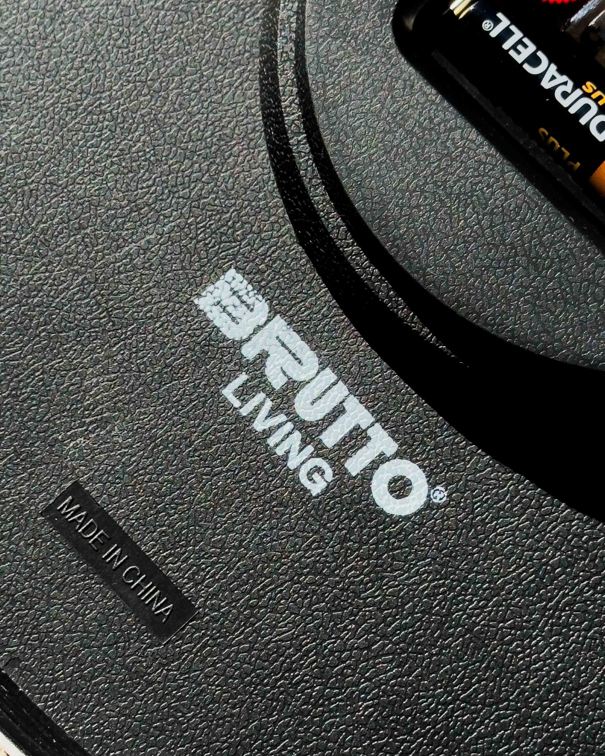 Close-up of a black textured surface with the white Brutto Studio logo, a MADE IN CHINA label, and part of a Duracell battery—perfect for your Brutto Studio Hurry Up! Wall Clock - Multicolour featuring silent quartz movement.
