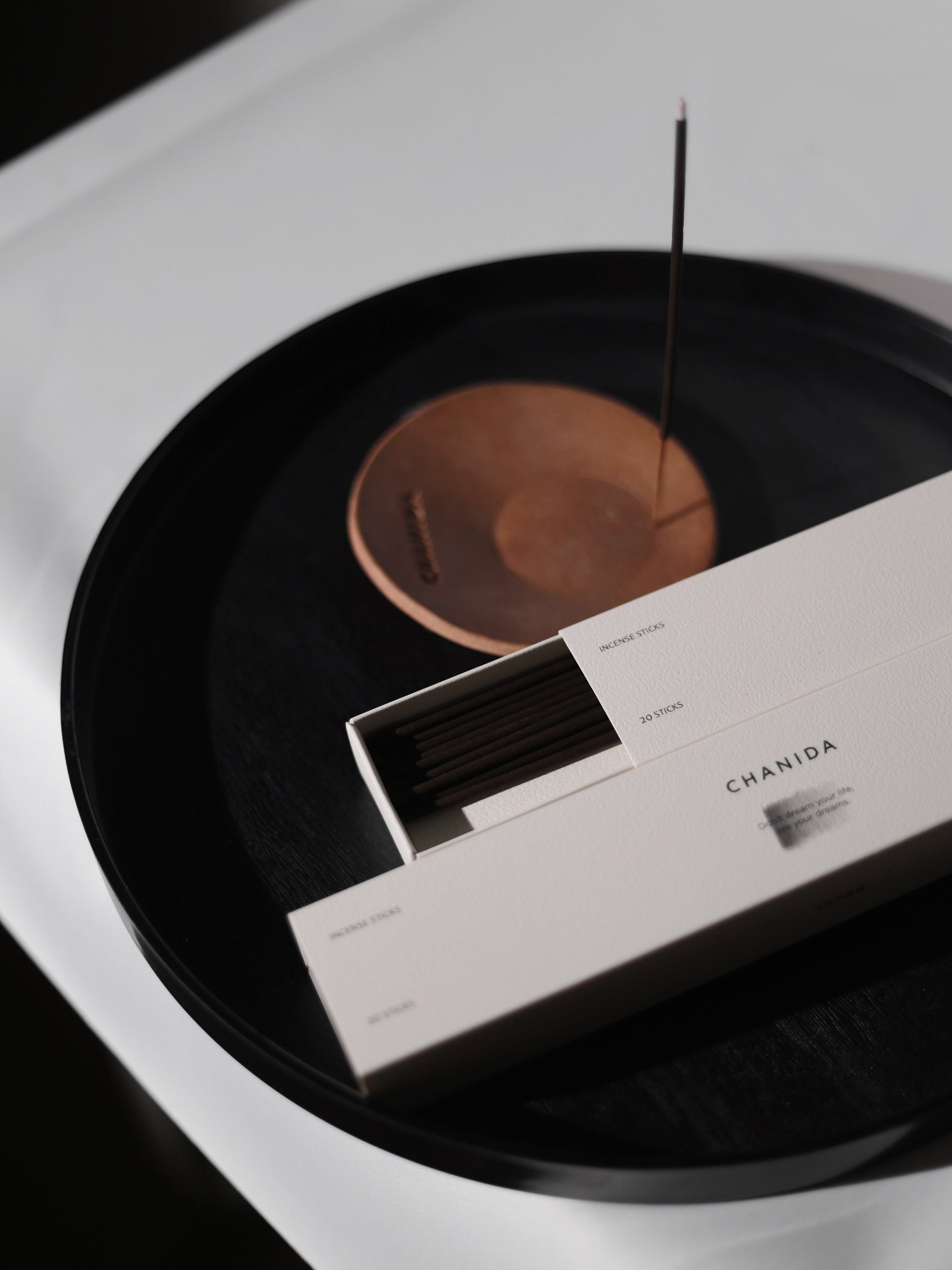An open box of CHANIDA Guaiacwood-Sandalwood Incense Sticks rests on a round black tray, with one stick burning in a brown holder nearby. The minimalistic setup features soft lighting and neutral tones for an elegant look.