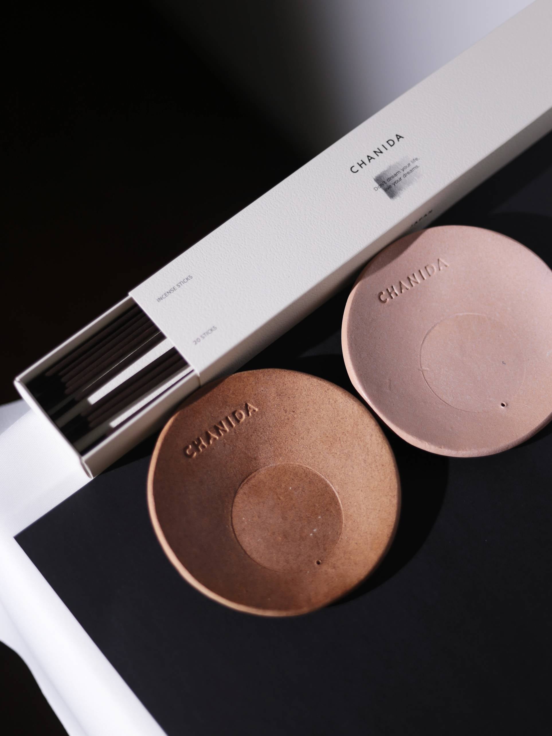 A white CHANIDA box of Japanese Green Tea-Vanilla Incense Sticks sits beside two round brown ceramic holders stamped CHANIDA, all arranged on a black and white surface, evoking an elegant Japanese ambiance.
