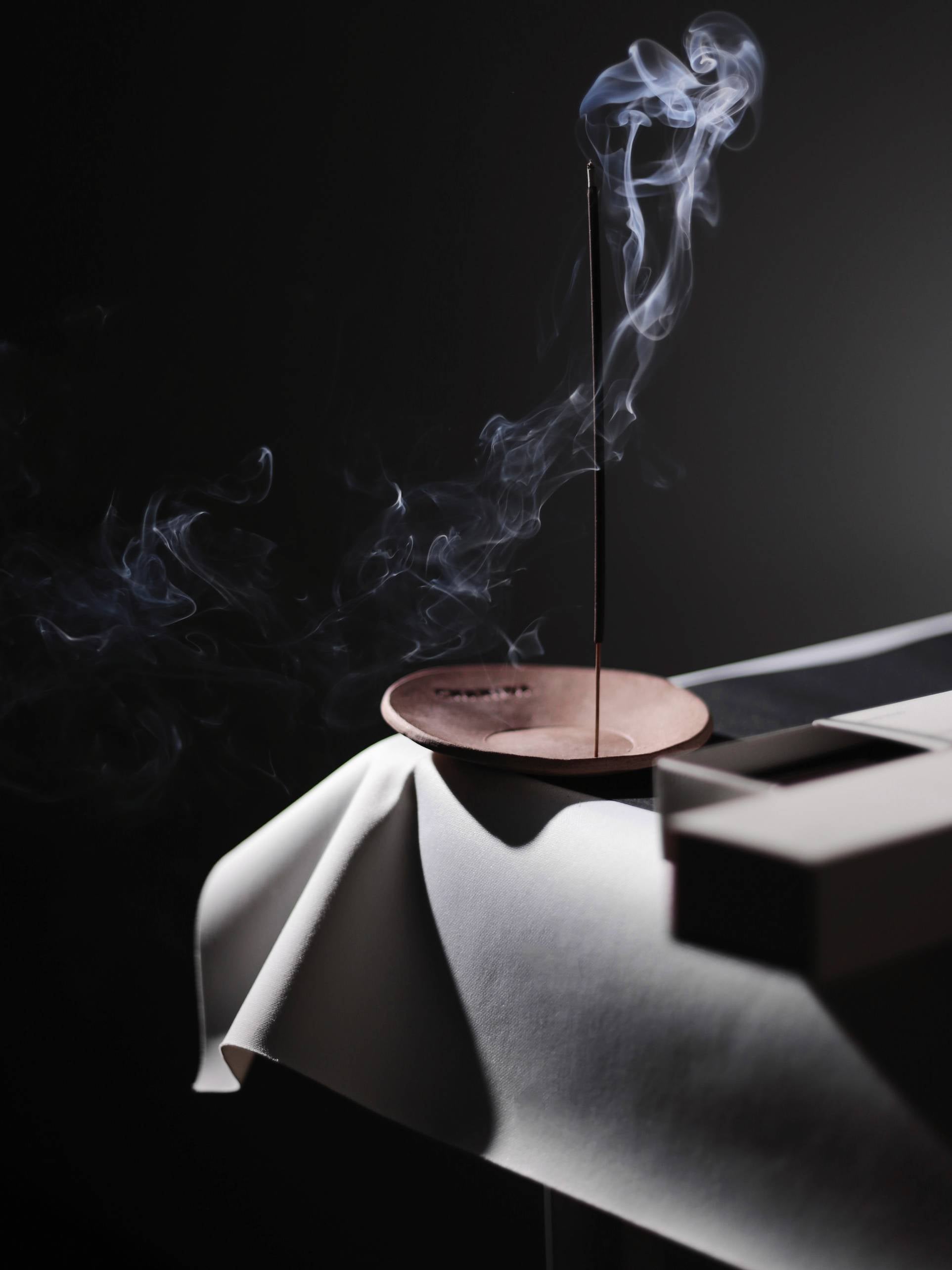 A lit CHANIDA Japanese Green Tea-Vanilla Incense Stick in a round holder rests on draped white cloth, smoke curling upward against a dark backdrop. A partially visible CHANIDA box sits nearby.