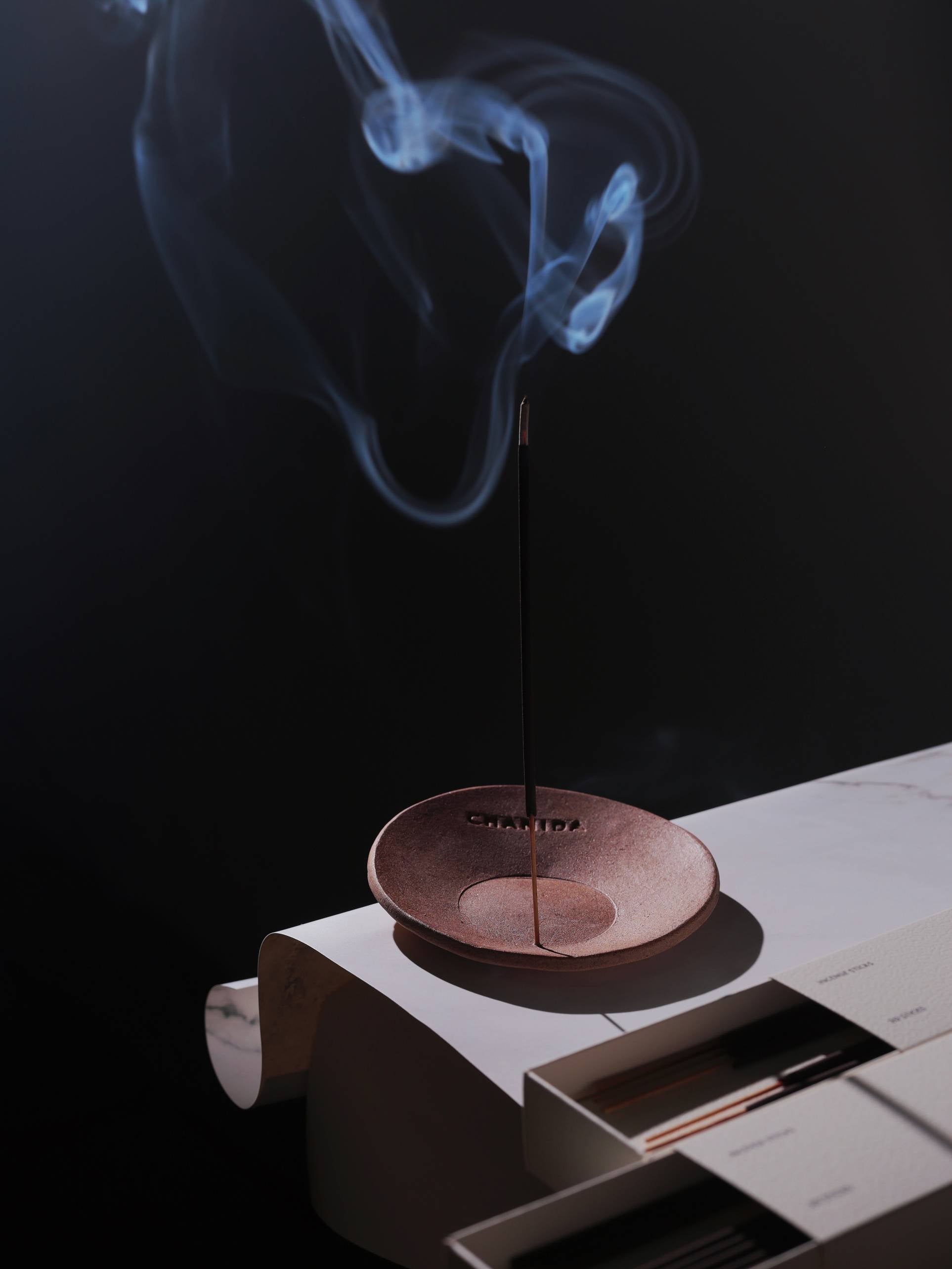 A CHANIDA Lavender-Musk Incense Stick burns in a small dish on a white surface beside a closed book, releasing curling smoke against a dark background.
