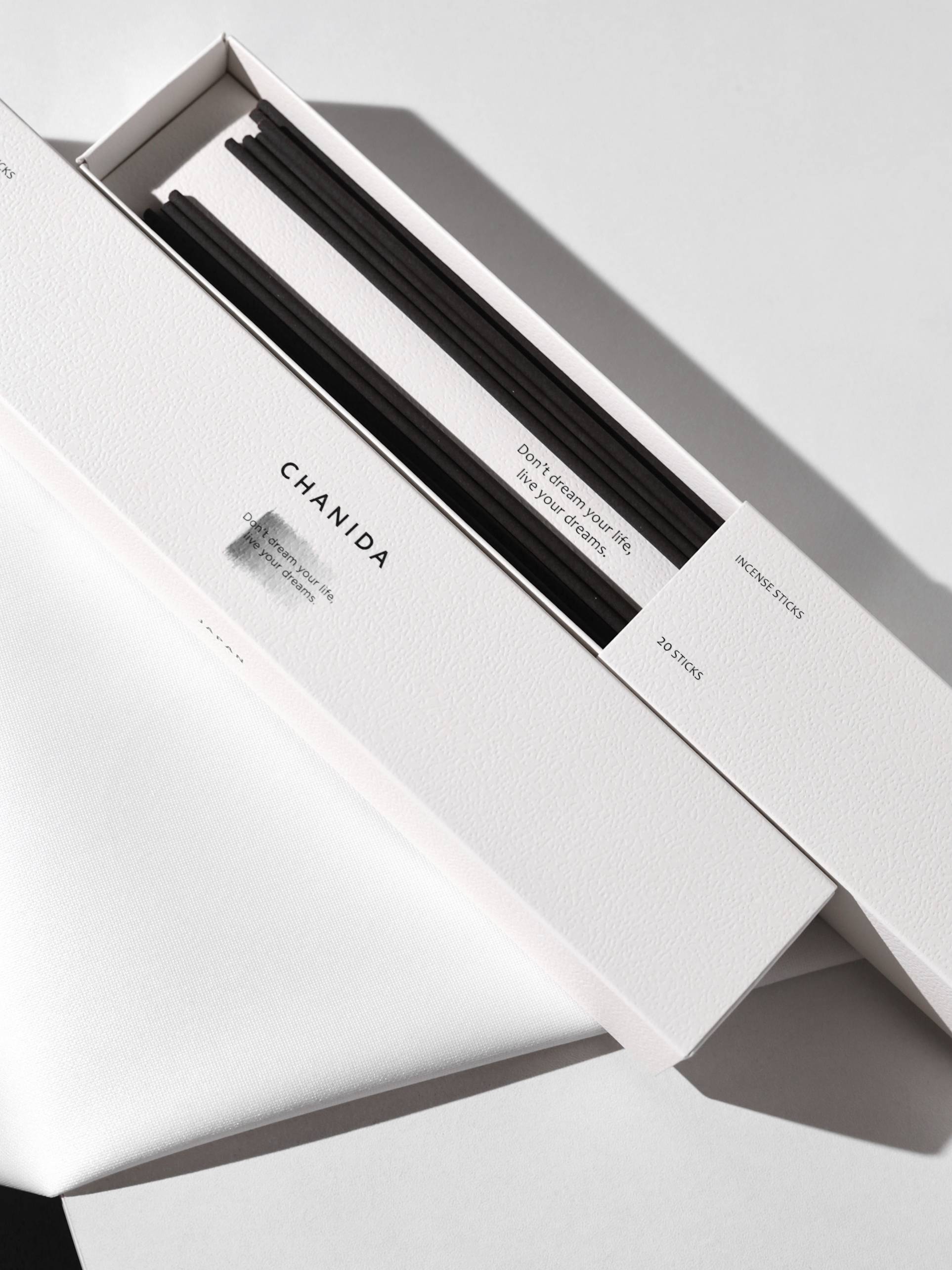 A white CHANIDA box labeled White Tea-Osmanthus Incense Sticks holds black incense sticks. The lid reads: Dont chase the bliss. Enjoy the calm, complementing the minimalist design of shadow and light.