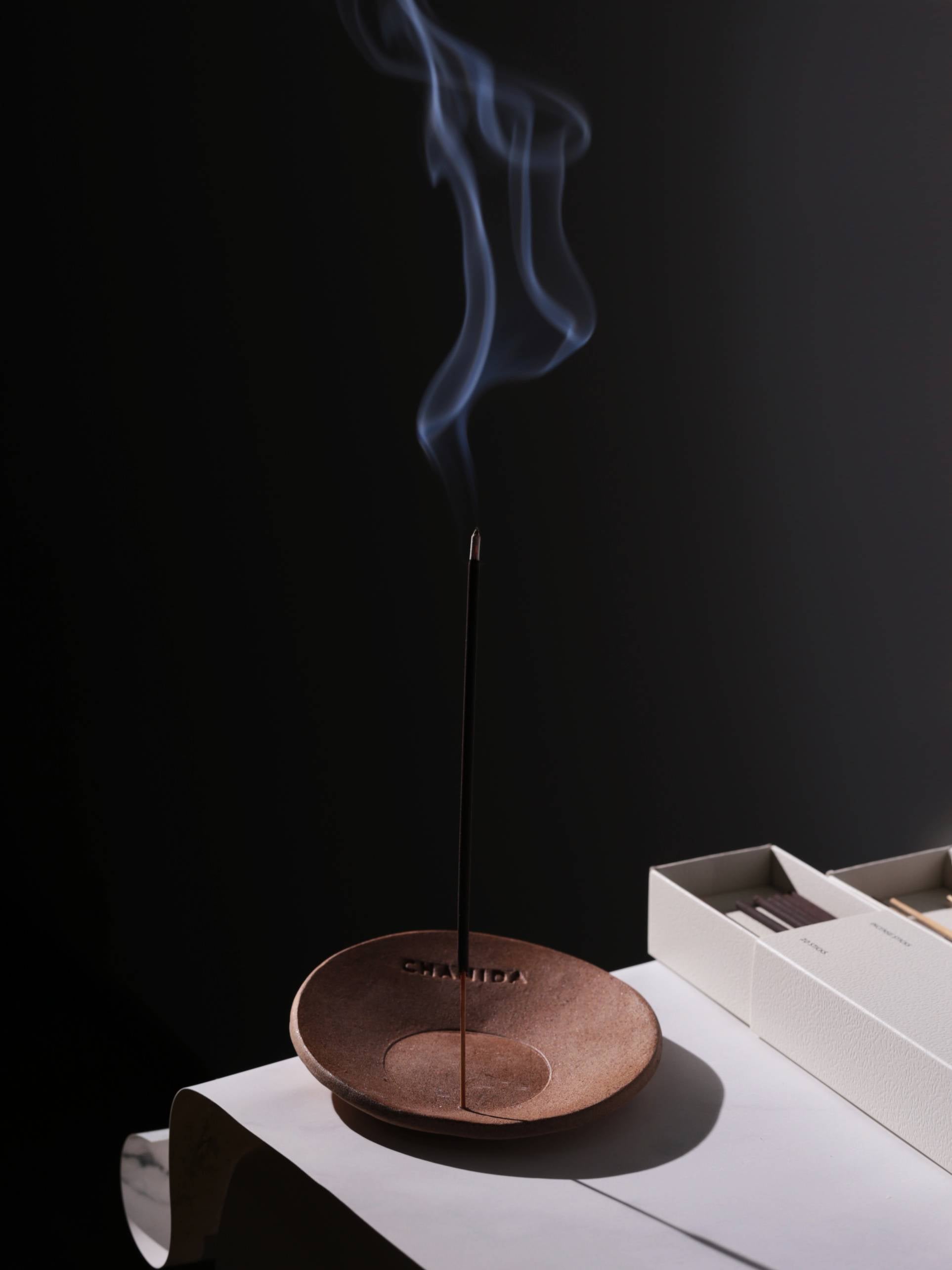A CHANIDA White Tea-Osmanthus Incense Stick rests in a round brown holder, gently releasing fragrant smoke on a white surface next to its beige box, all set against a dark backdrop.