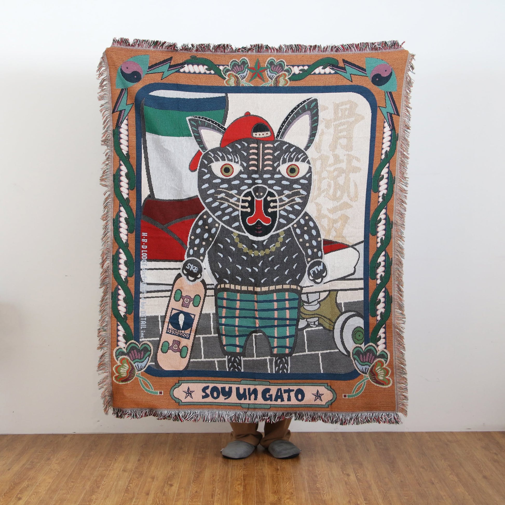A person is holding the DETAIL Black Cat Oax Throwket by DETAIL, a vibrant multicolour blanket featuring a cartoon cat in a red cap with a skateboard, SOY UN GATO at the bottom, and decorative border patterns.