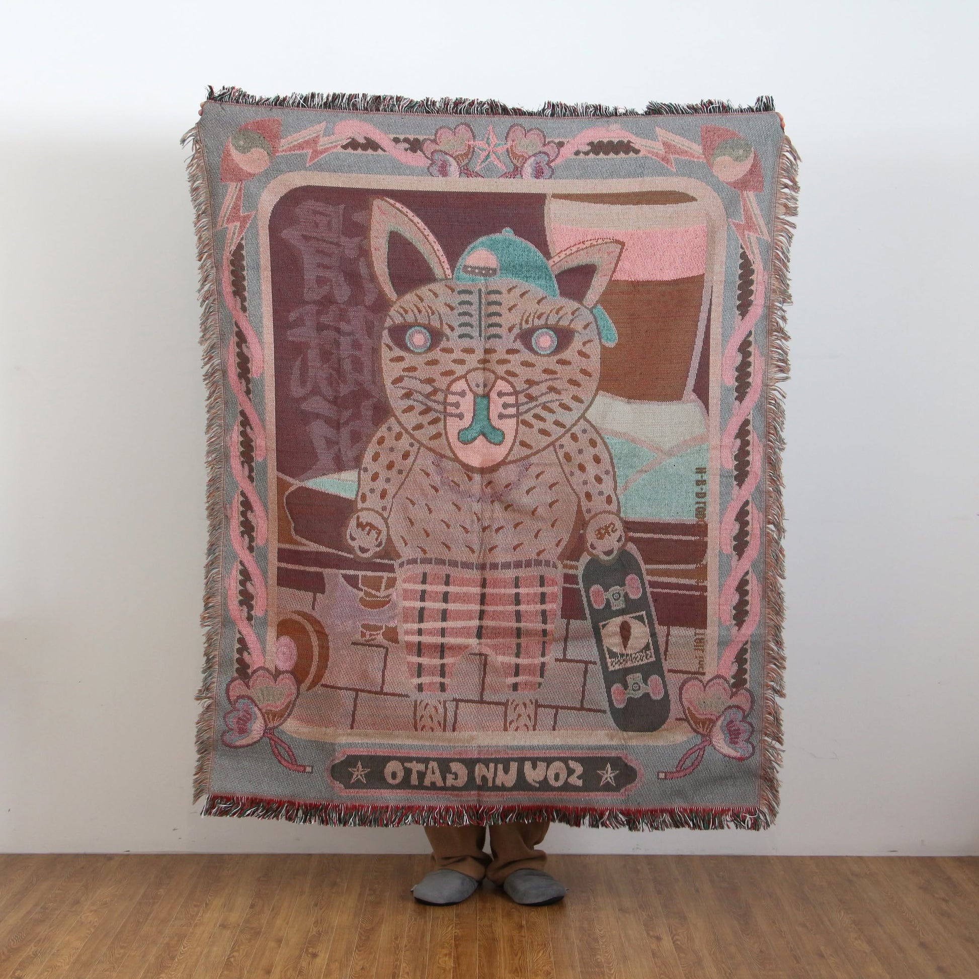 A person stands behind the DETAIL Black Cat Oax Throwket by DETAIL, featuring a cartoon cat with a hat and skateboard. Only their feet are visible below the multicolour throw blanket with decorative borders and mirrored text.