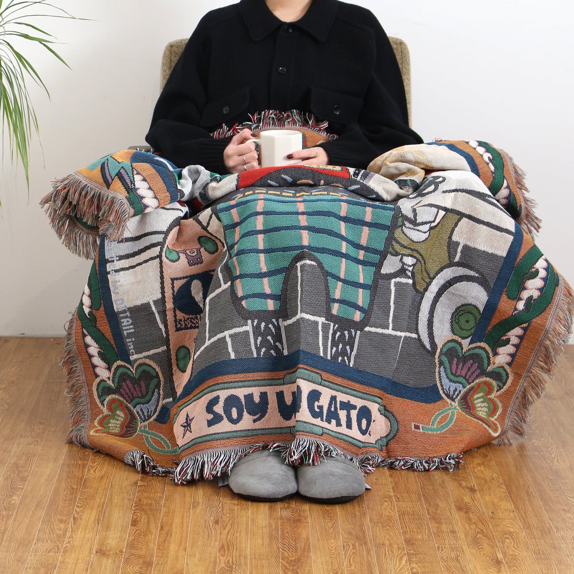 Wrapped in the DETAIL Cat Oax Throwket by DETAIL, featuring folk art cat designs and SOY UN GATO, a person sits cozily with a white mug and grey slippers, enjoying this charming, colorful cat-themed accessory.