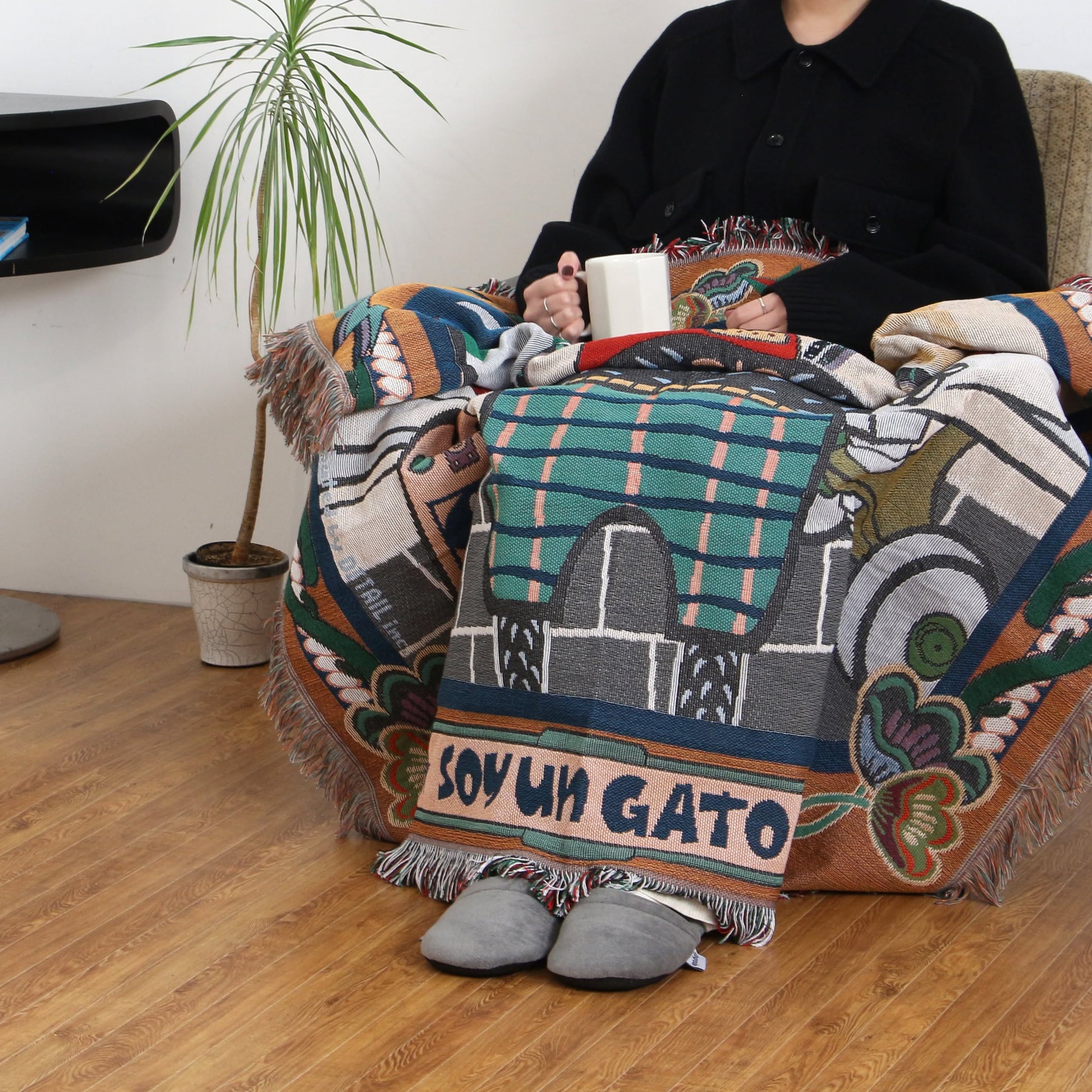 A person relaxes in an armchair with the DETAIL Cat Oax Throwket by DETAIL, a vibrant cat-themed blanket featuring SOY UN GATO, holding a white mug and wearing gray slippers beside a potted plant on a wooden floor.