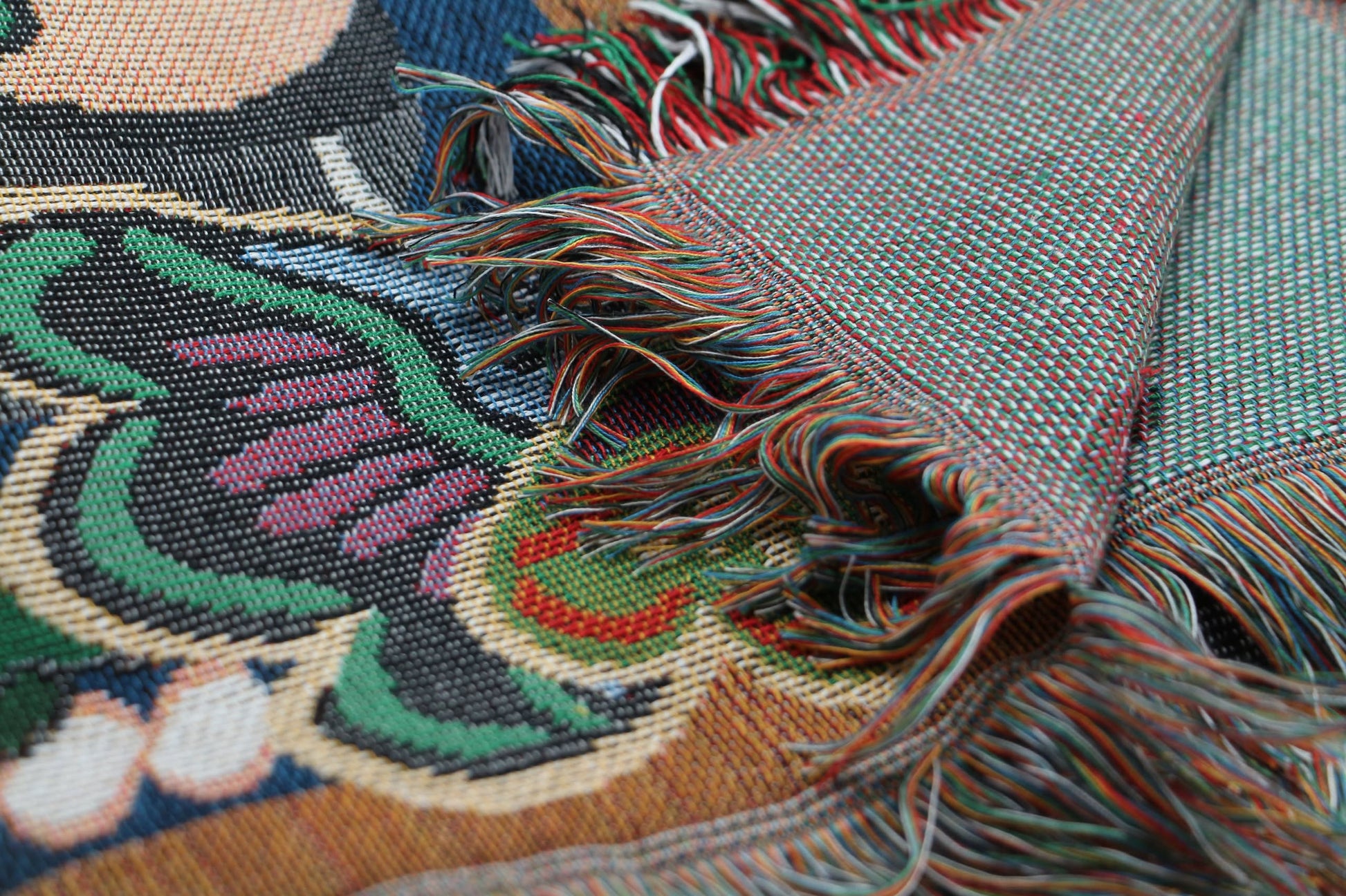 A close-up of the DETAIL Black Cat Oax Throwket by DETAIL highlights its vibrant artisanal design, featuring green, red, white, and black threads and detailed fringes with the intricate reverse side visible.