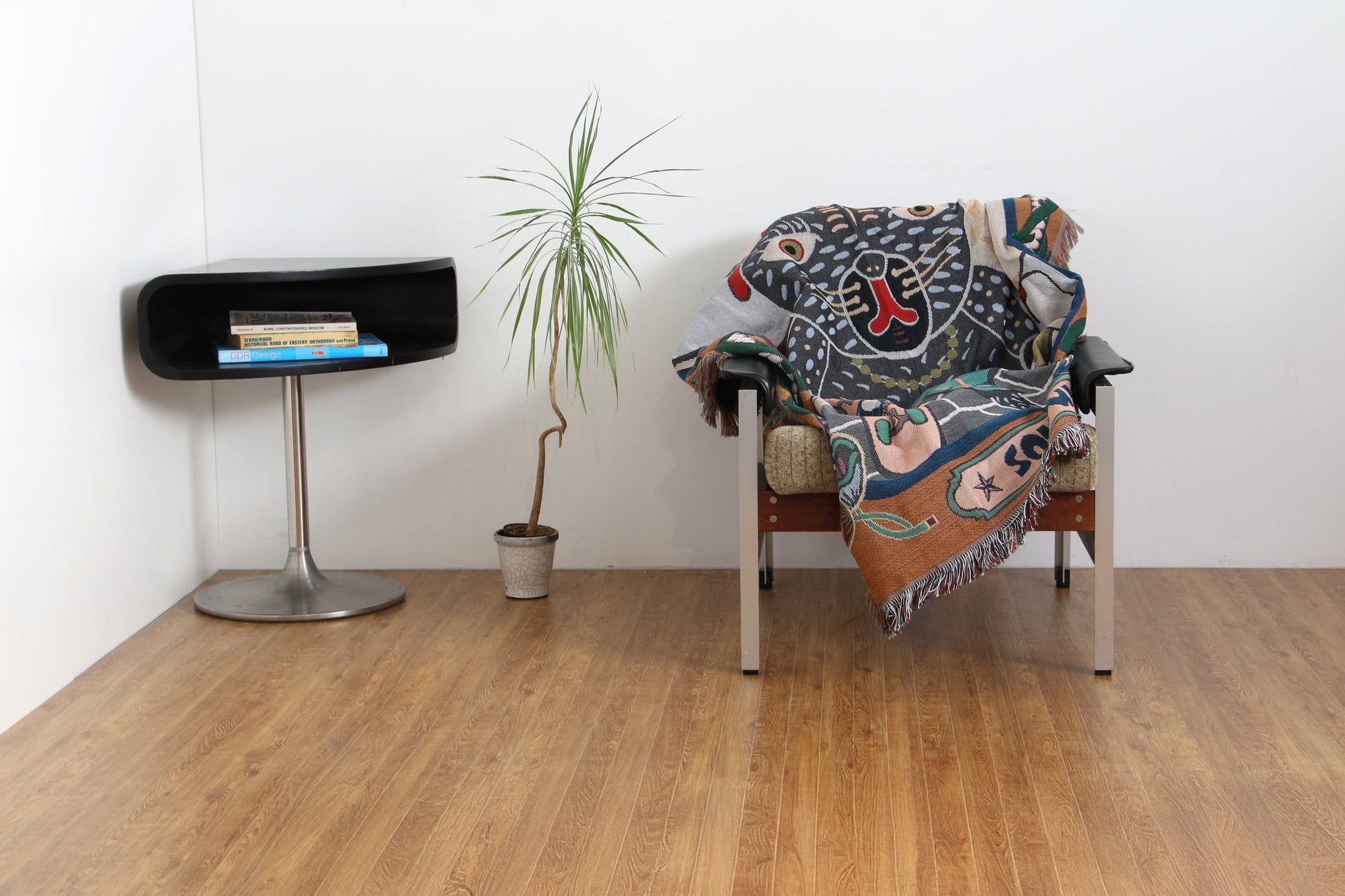 A cozy armchair with the DETAIL Black Cat Oax Throwket by DETAIL draped over it sits on a wooden floor beside a small potted plant and a modern side table with books, set against a plain white wall.
