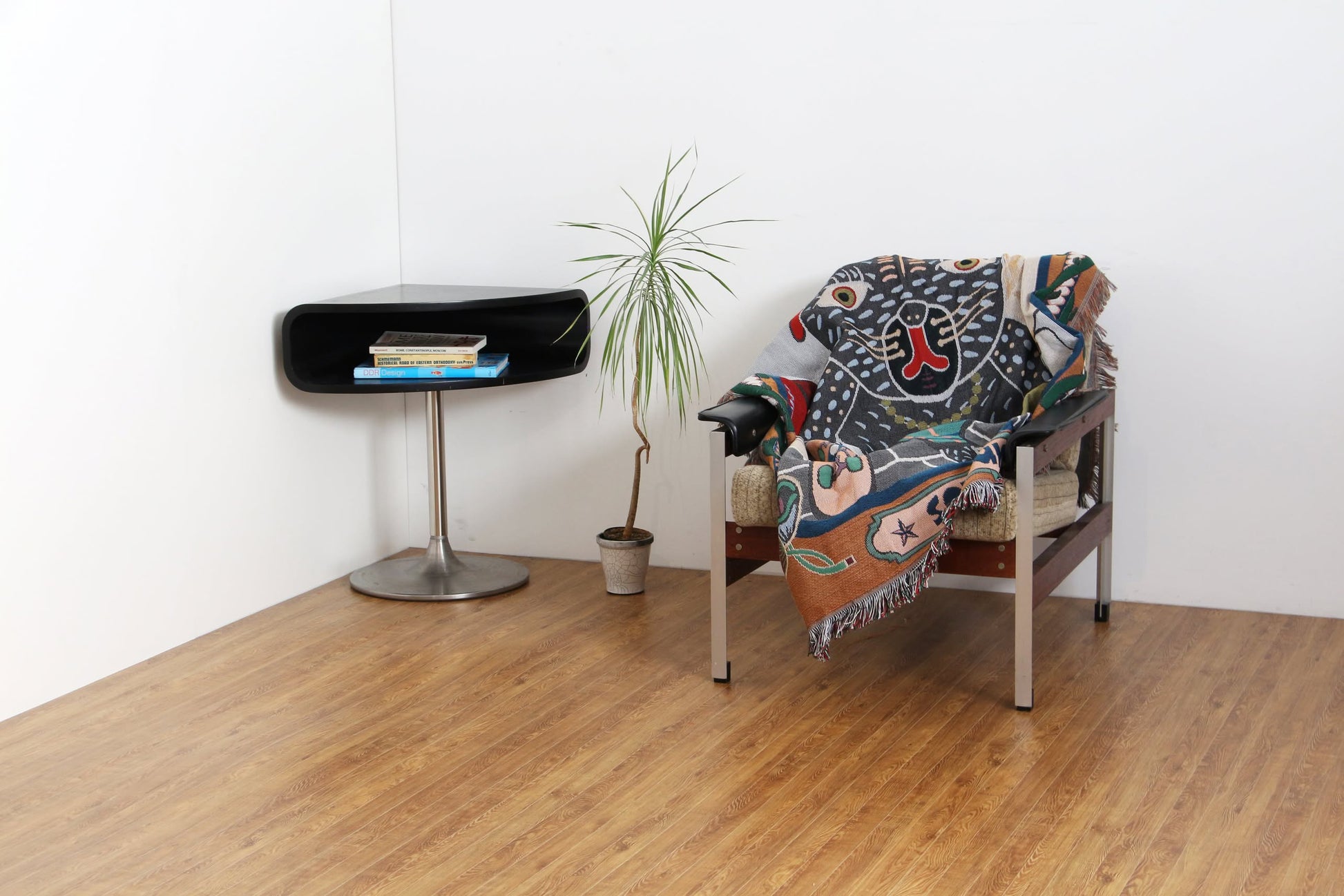 A modern, minimalist room features a chair draped with the DETAIL Black Cat Oax Throwket by DETAIL, a small potted plant, and a black side table with books on wooden floors against white walls.