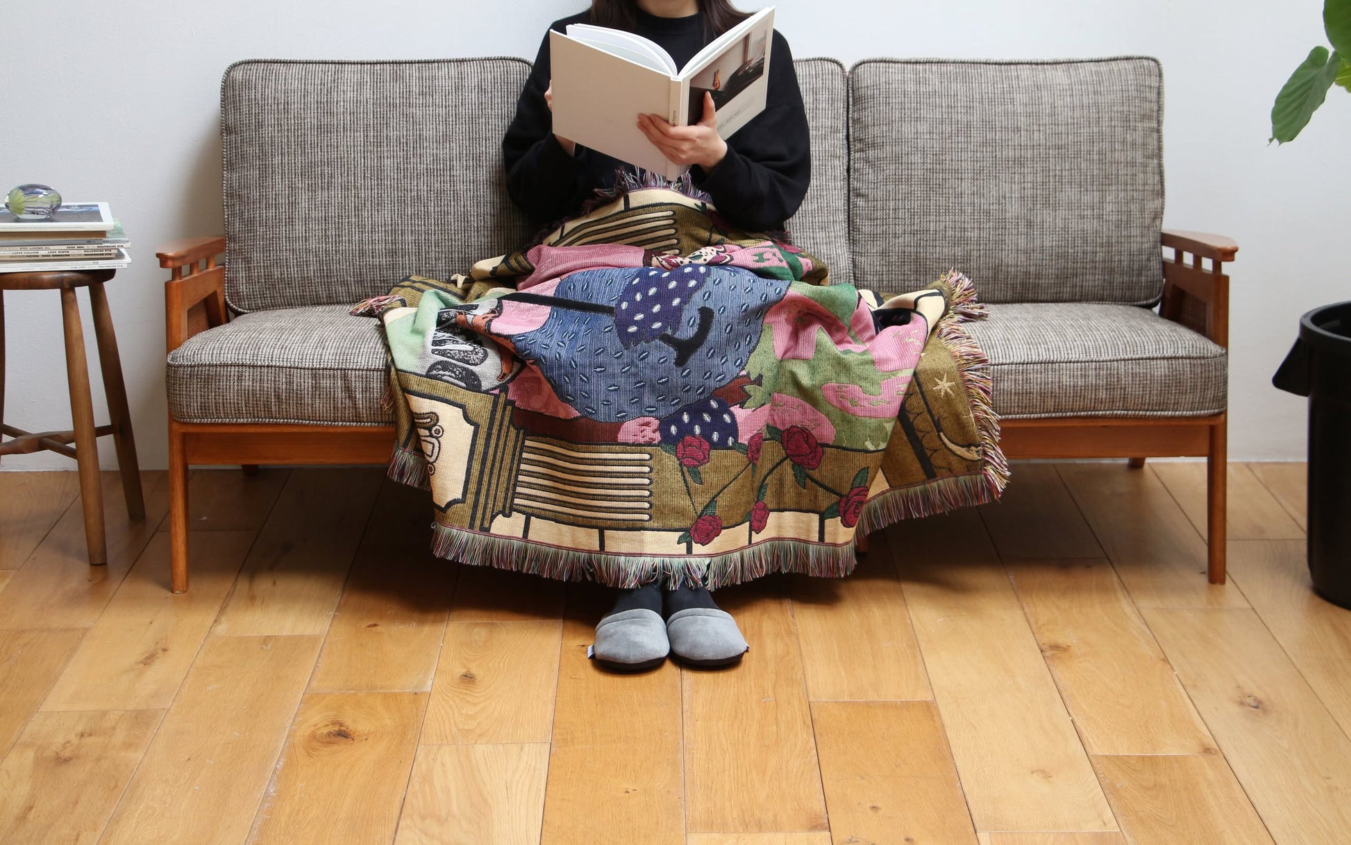 A person sits on a gray sofa with the DETAIL Dodo Oax Throwket by DETAIL draped over their lap, reading a book. Only the book and lower half of their body are visible; their face is out of frame.