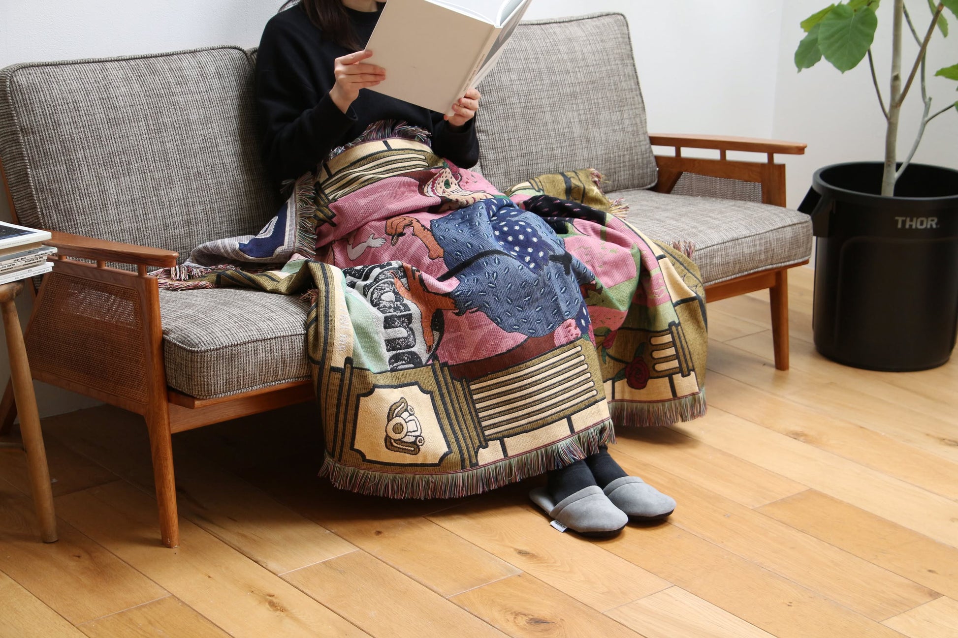 Wrapped in the vibrant DETAIL Dodo Oax Throwket by DETAIL, a person wearing dark clothes and gray slippers relaxes on a sofa reading, surrounded by wooden floors, a potted plant, and Oaxaca-inspired décor in a cozy living room.