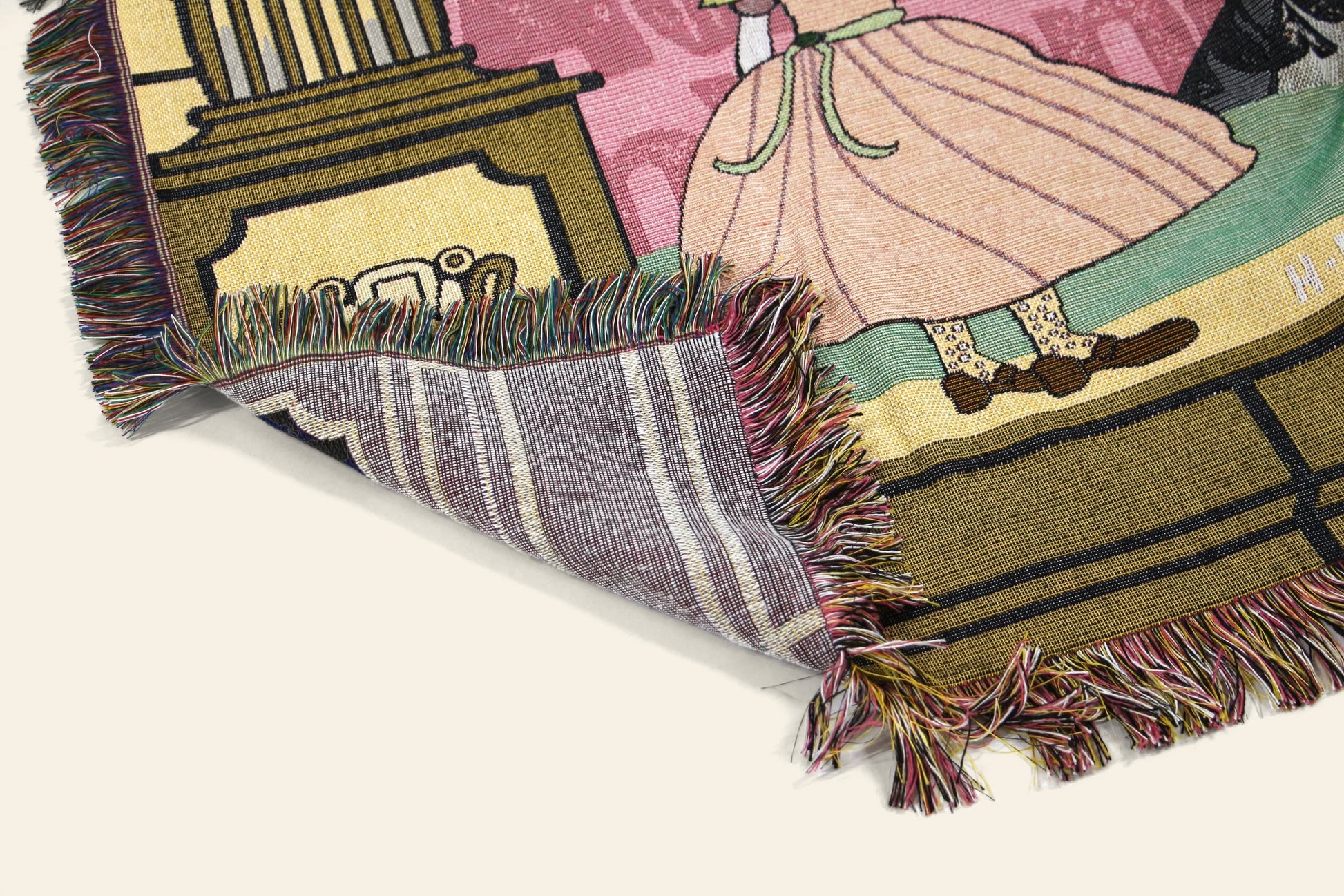 The DETAIL Dodo Oax Throwket by DETAIL is a colorful folk art throw with multicolored fringes, showcasing a person in a pink dress and brown shoes by a column. Its folded corner reveals the Oaxaca-inspired underside design.