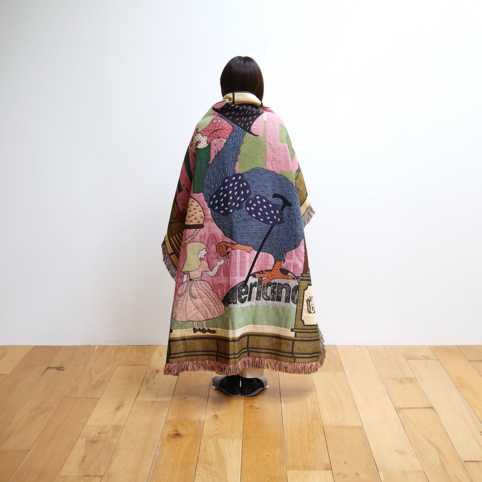 A person stands on a wooden floor facing a white wall, draped in the DETAIL Dodo Oax Throwket by DETAIL, a colorful folk art blanket featuring large illustrations of birds and a person in a dress, with American visible.