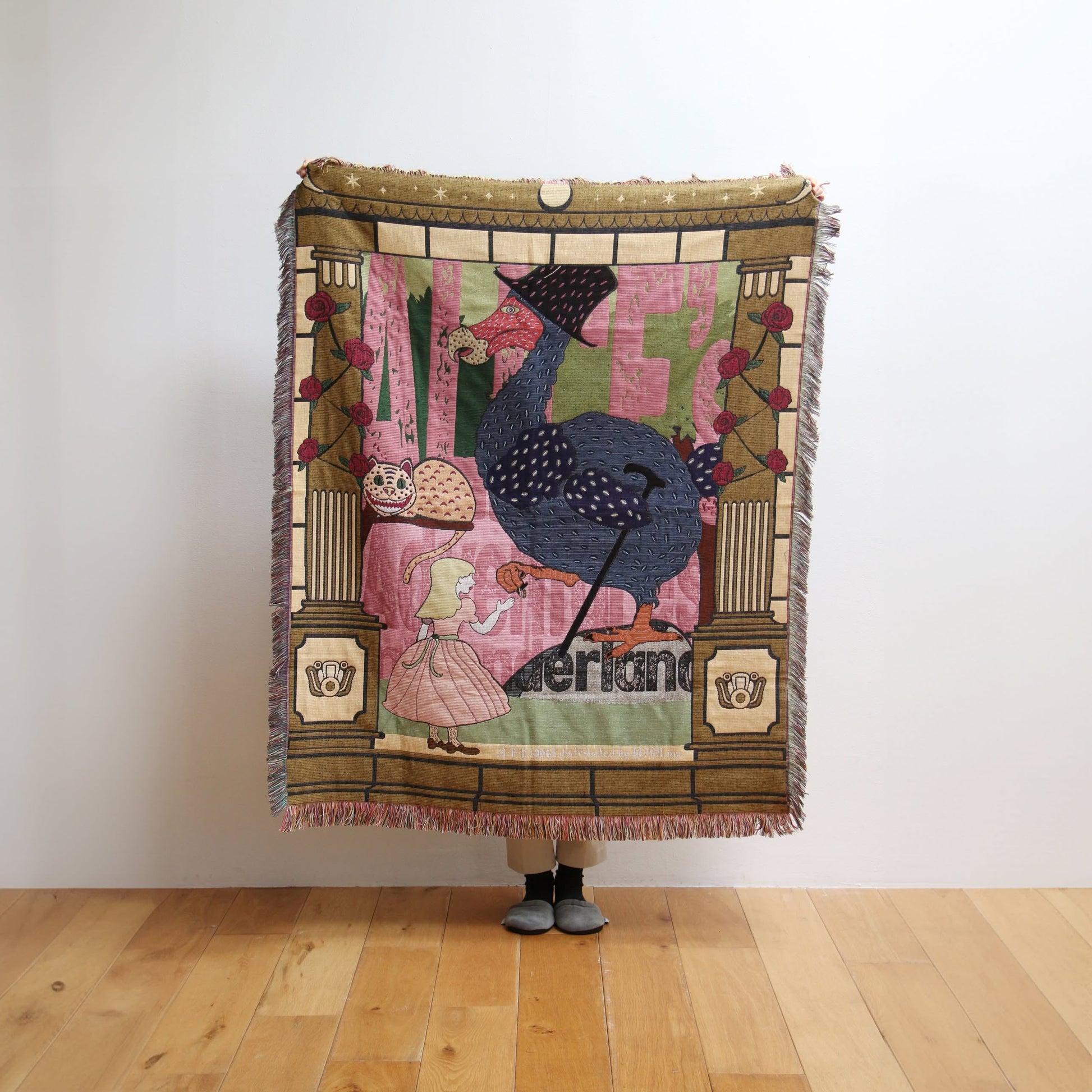 A person stands on a wooden floor holding the DETAIL Dodo Oax Throwket by DETAIL, featuring an illustrated young girl, a large blue bird in a top hat, and whimsical elements on a theatrical backdrop. Only their shoes are visible.