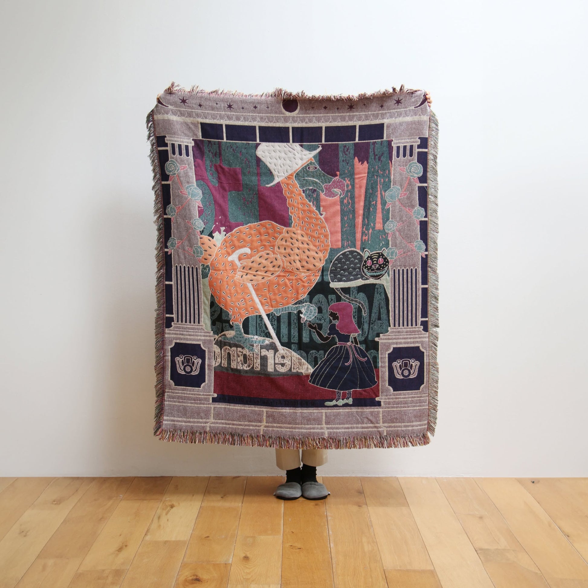 A person stands on a wooden floor holding up the DETAIL Dodo Oax Throwket by DETAIL, featuring a colorful folk art illustration of a large bird in a bonnet and a girl in a dress; only the person’s shoes are visible below the throw blanket.