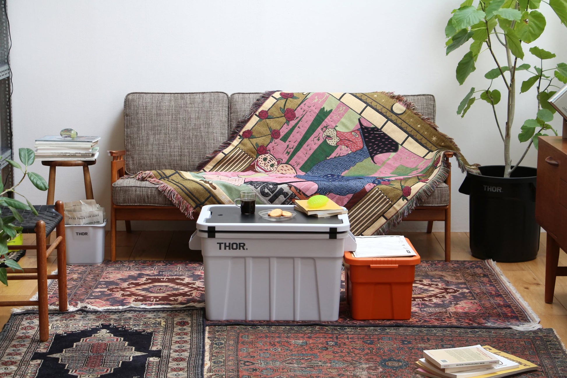 A cozy living room features the DETAIL Dodo Oax Throwket by DETAIL on a vintage sofa, white and orange storage bins as a coffee table, books, houseplants, and Oaxaca-inspired patterned rugs on a wooden floor.