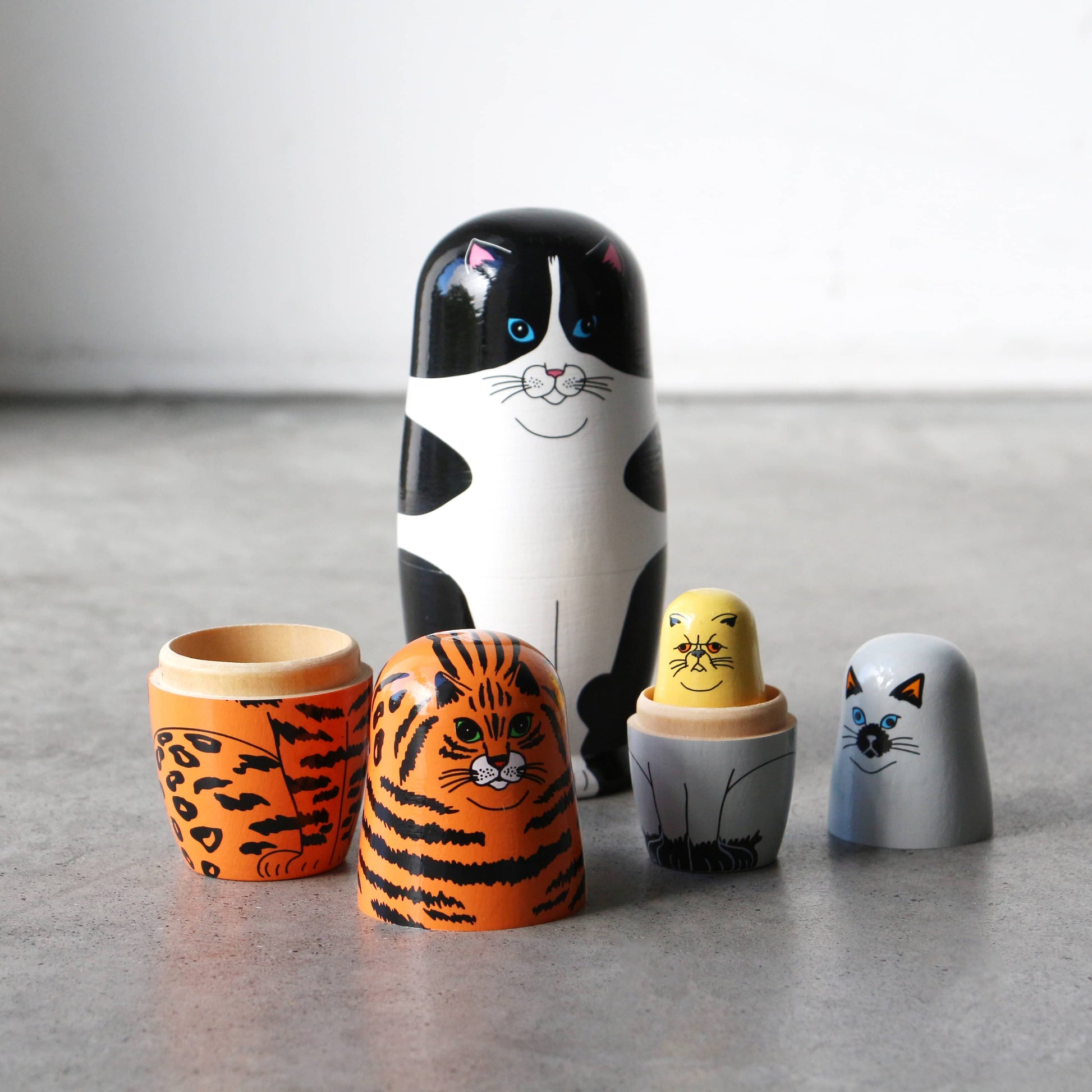 DETAIL Feline 5 Nesting Dolls by DETAIL feature charming wooden cats and tigers, some opened to reveal smaller dolls, arranged on a gray surface. Perfect for cat lovers, they stand out beautifully against the plain light background.