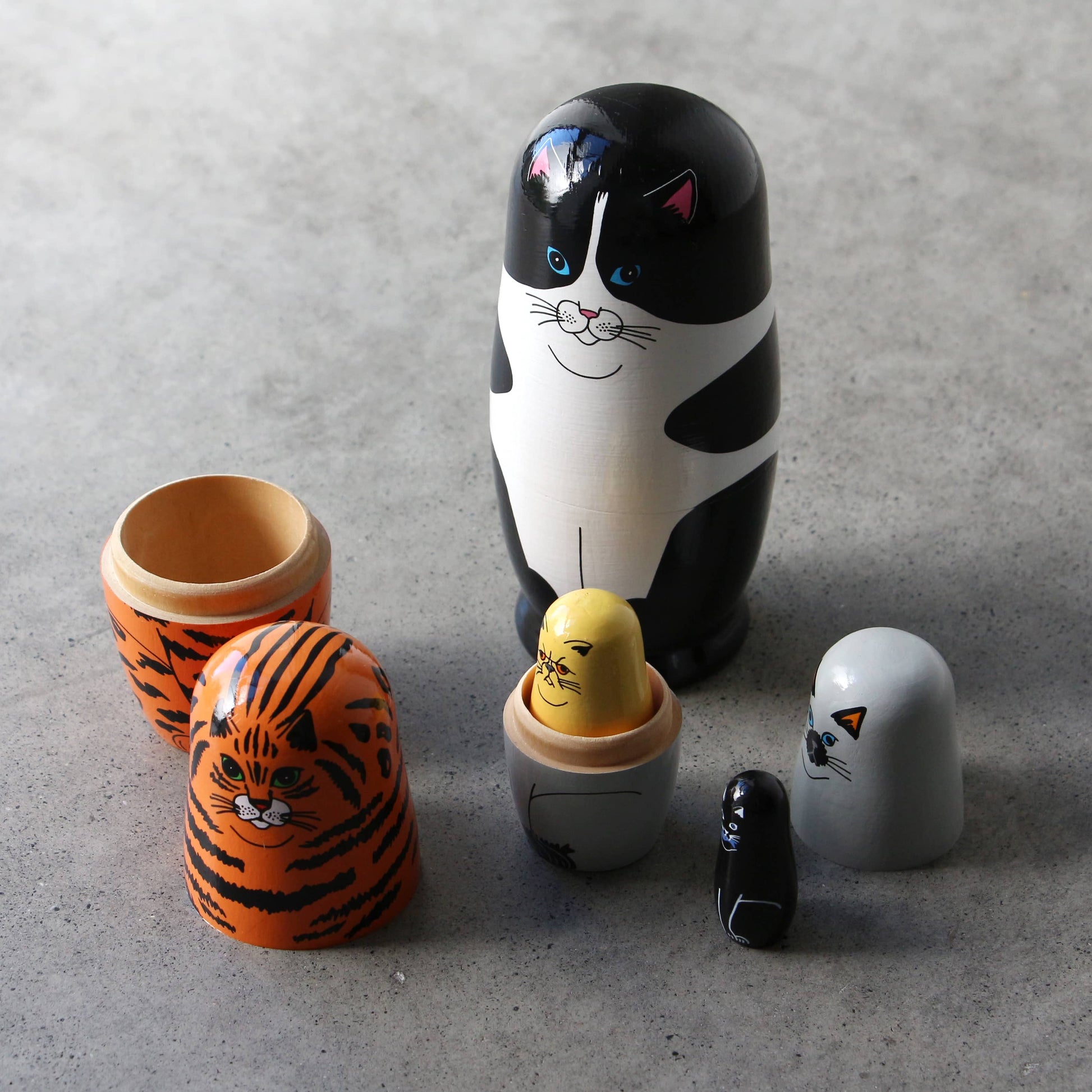 Five DETAIL Feline 5 Nesting Dolls by DETAIL, featuring cat designs in different colors, are shown on a gray surface with some opened to reveal smaller dolls inside—an ideal gift for cat lovers.