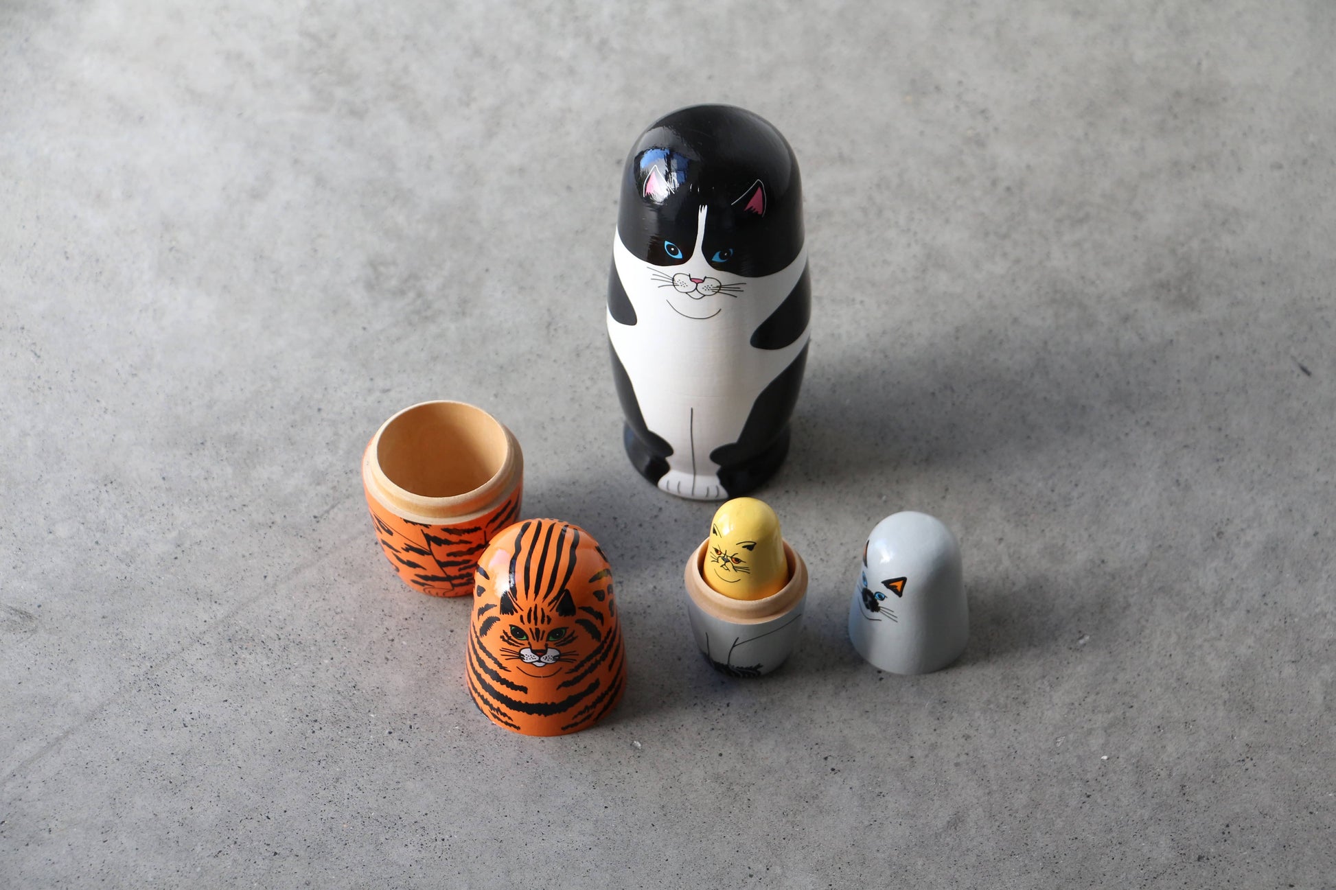 The DETAIL Feline 5 Nesting Dolls by DETAIL features five wooden dolls—a black-and-white cat, tiger, yellow chick, grey penguin, and more—displayed opened on a gray surface; ideal for cat lovers and collectors.
