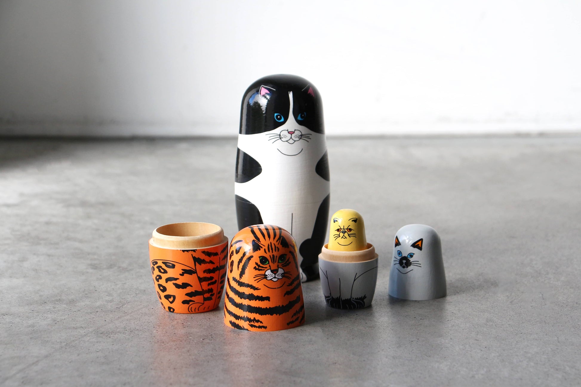 The DETAIL Feline 5 Nesting Dolls by DETAIL feature wooden dolls in various sizes, each painted to resemble a different cat. Arranged on a gray surface, these nesting dolls are perfect for cat lovers with their unique expressions and patterns.