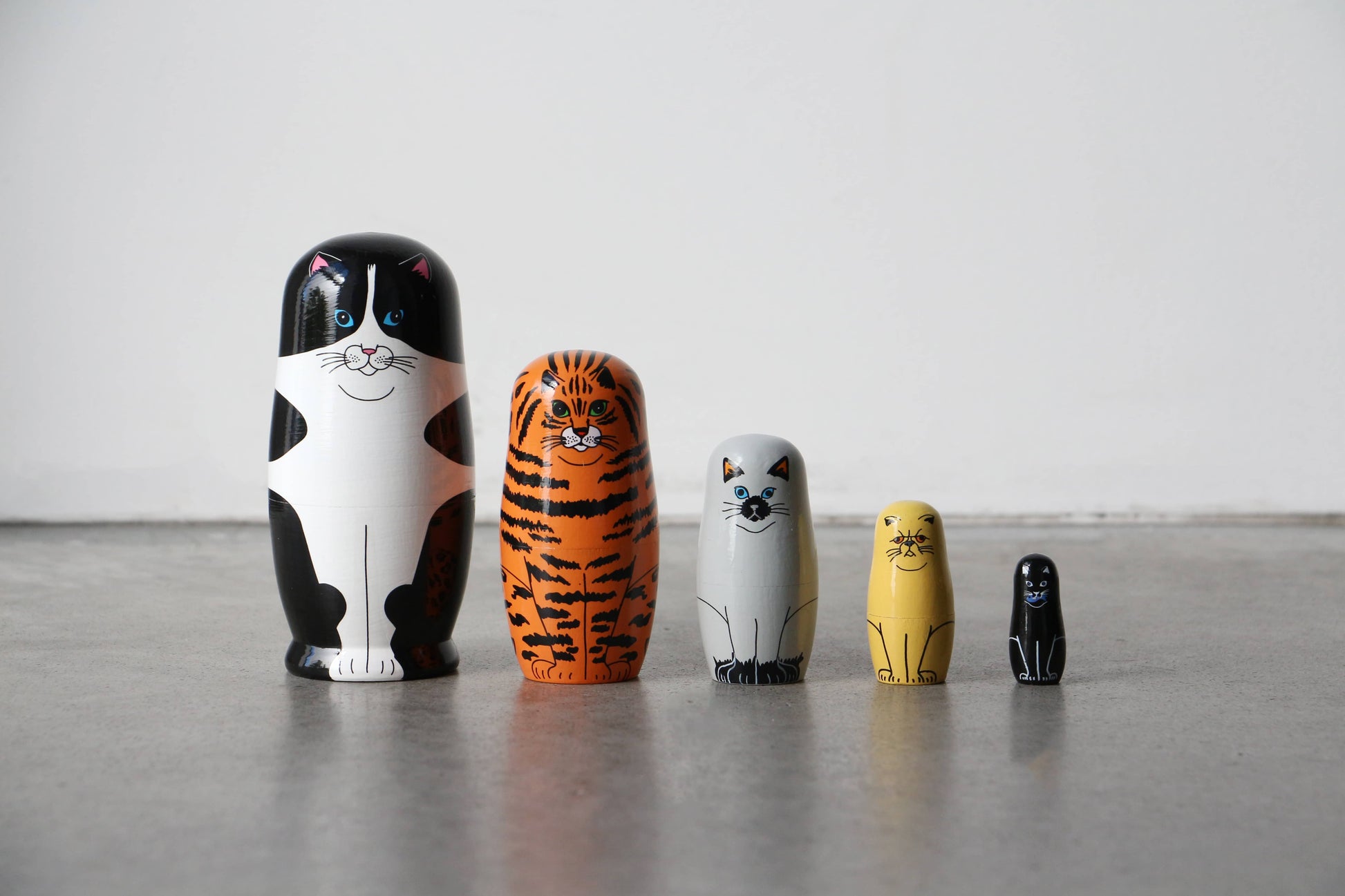 The DETAIL Feline 5 Nesting Dolls by DETAIL features five wooden cat-shaped dolls of different sizes and colors, arranged on a smooth gray floor against a white wall. Each doll is painted to resemble a unique type of cat—perfect for cat lovers.
