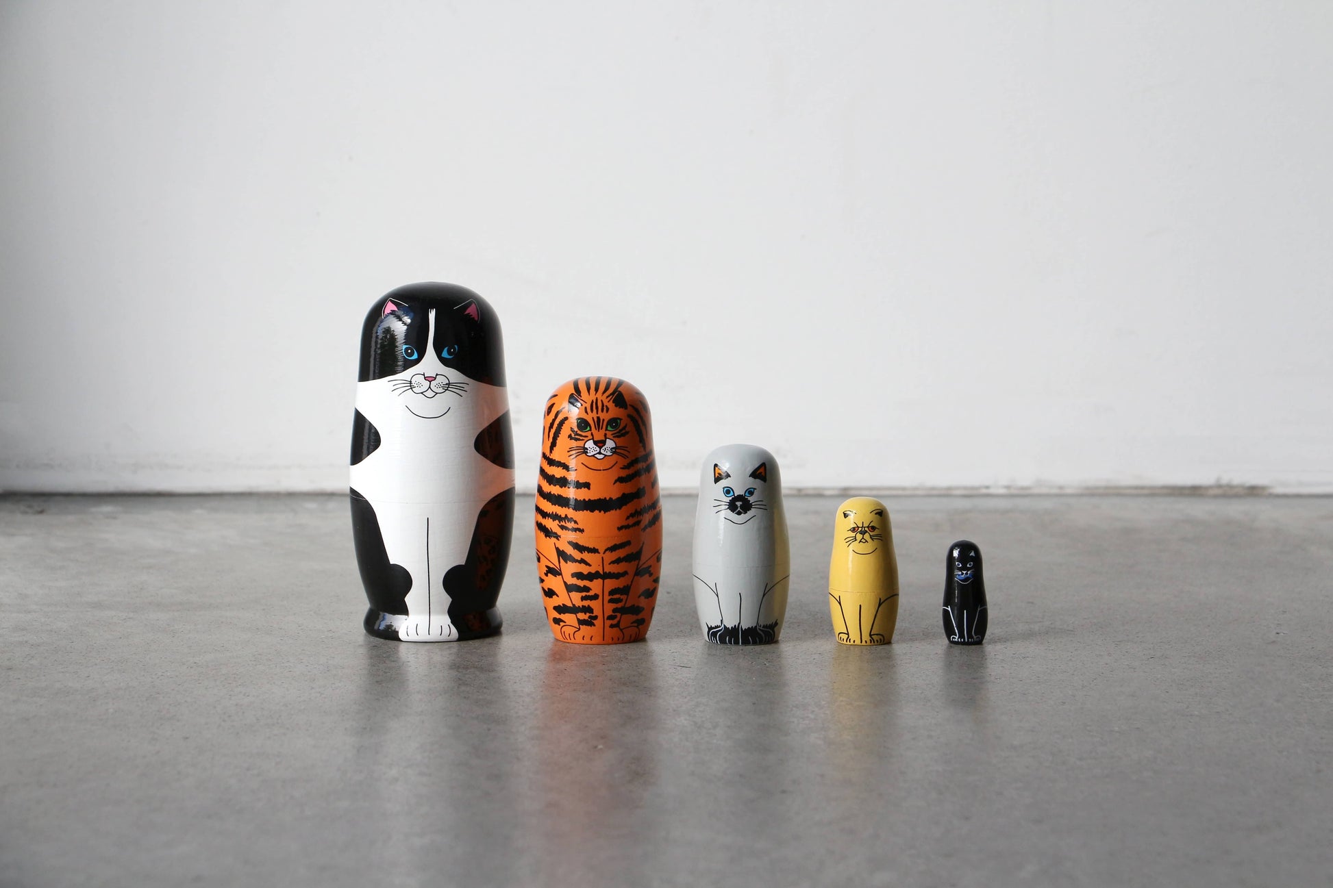 DETAIL Feline 5 Nesting Dolls by DETAIL, featuring five cat-themed dolls in different sizes and colors, are displayed in a row on a gray floor against a plain white wall—an ideal gift for cat lovers.