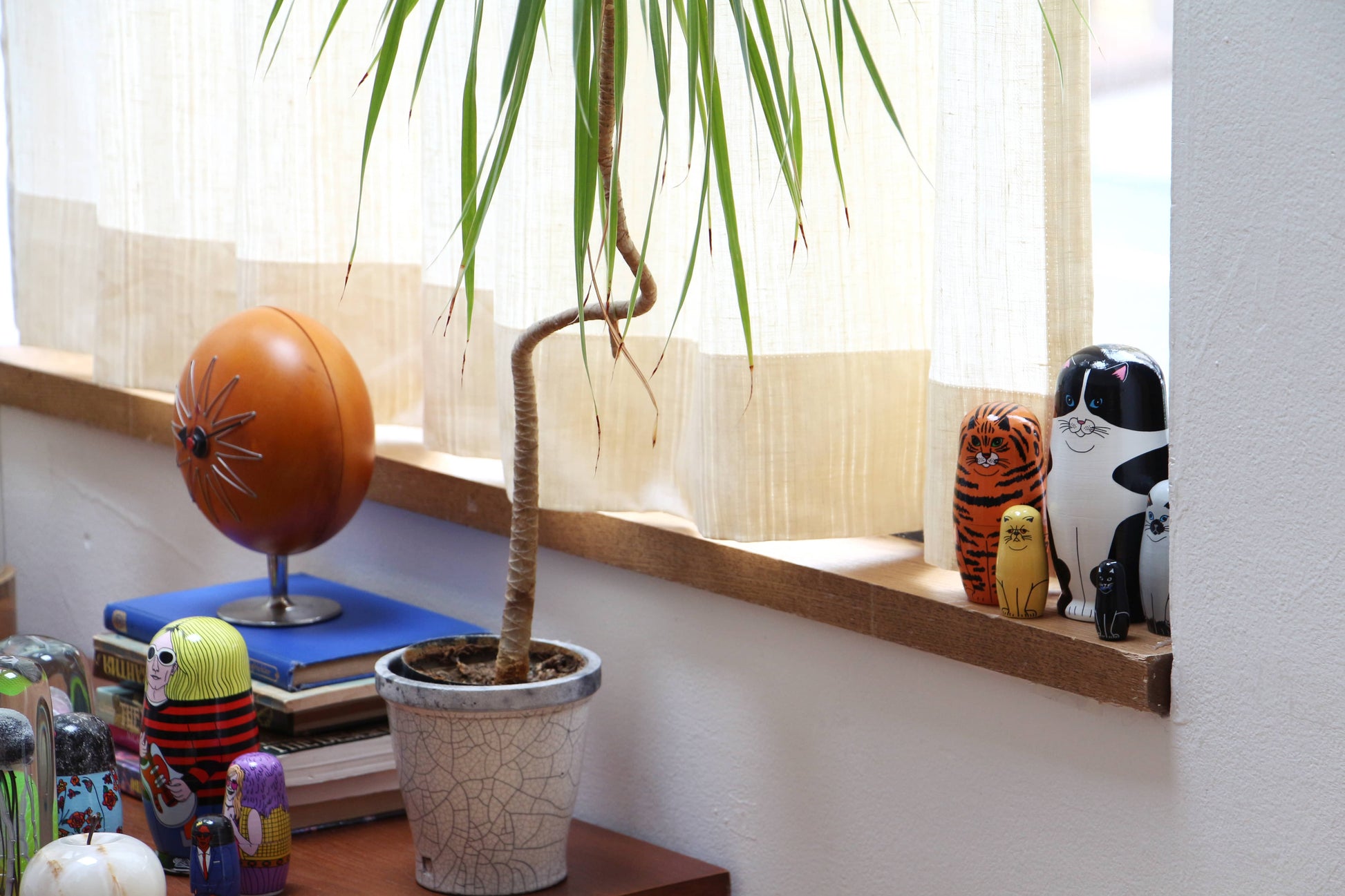 A potted plant with a twisted stem sits on a windowsill, surrounded by DETAIL Feline 5 Nesting Dolls from DETAIL, a tiger figurine, a black-and-white cat figurine—great for cat lovers—books, and an orange spherical lamp. Light filters through cream curtains.