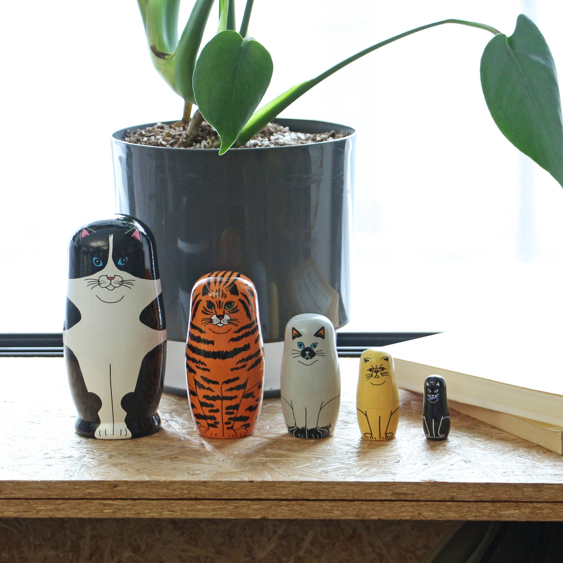 The DETAIL Feline 5 Nesting Dolls by DETAIL, in varying sizes, are arranged on wood near a gray potted plant and an open book with natural light—charming decor for cat lovers.