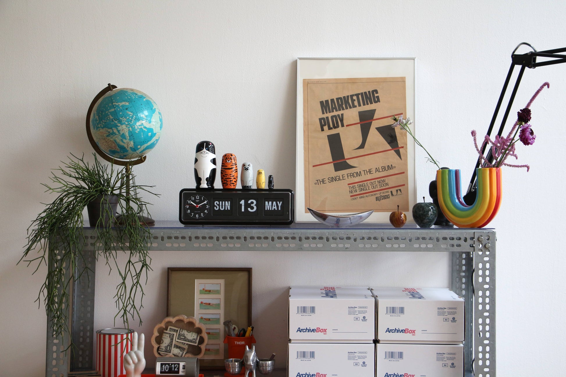 A metal shelf displays a globe, digital clock, potted plant, framed “MARKETING PLOY” poster, rainbow vase with flowers, stacked boxes, and DETAIL Feline 5 Nesting Dolls by DETAIL—ideal for cat lovers and decor enthusiasts.