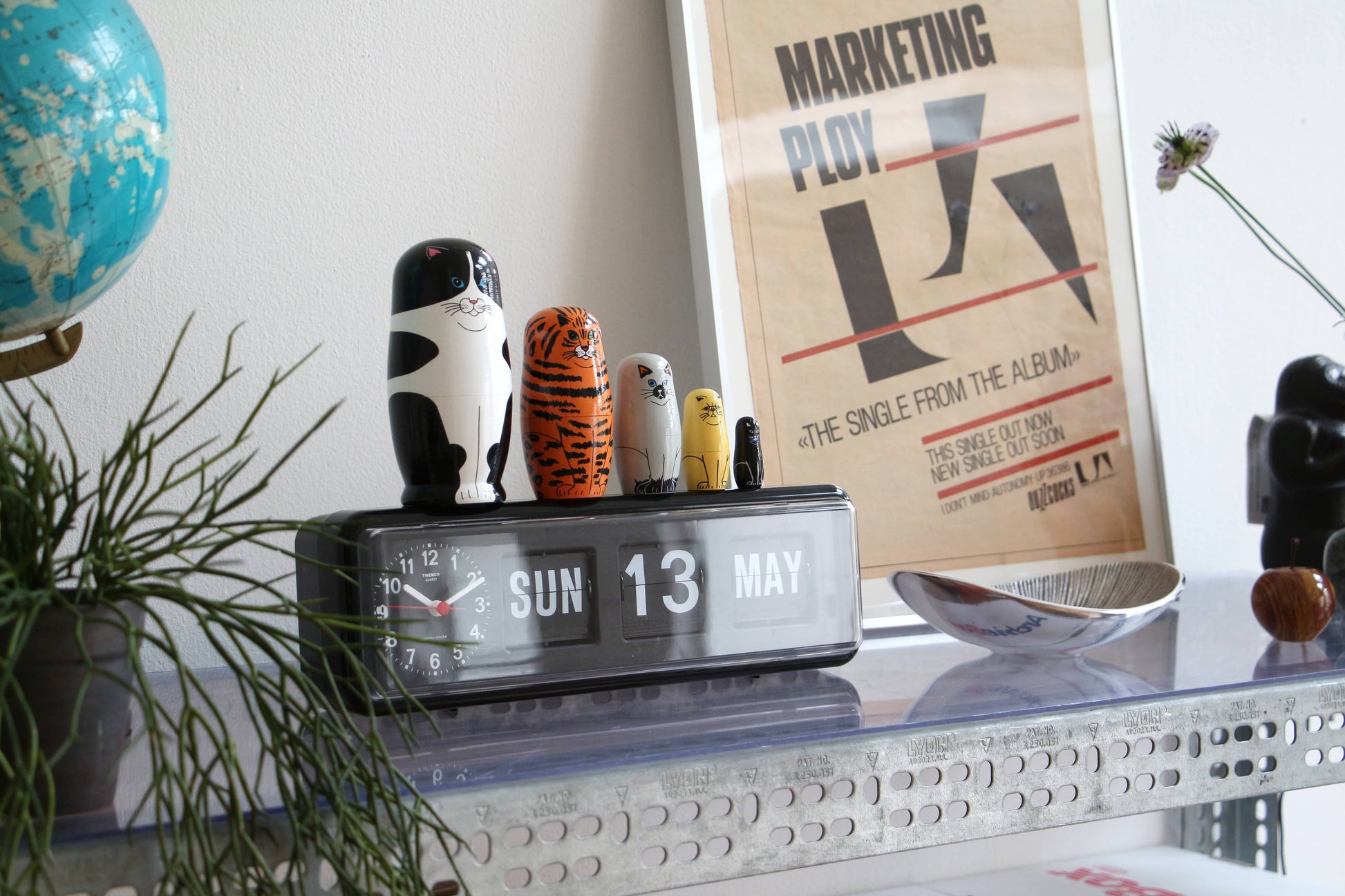 A metal shelf displays a flip clock set to “Sun 13 May,” the DETAIL Feline 5 Nesting Dolls by DETAIL, a globe, a modern spoon sculpture, a plant, and a framed marketing poster leaning against the wall.