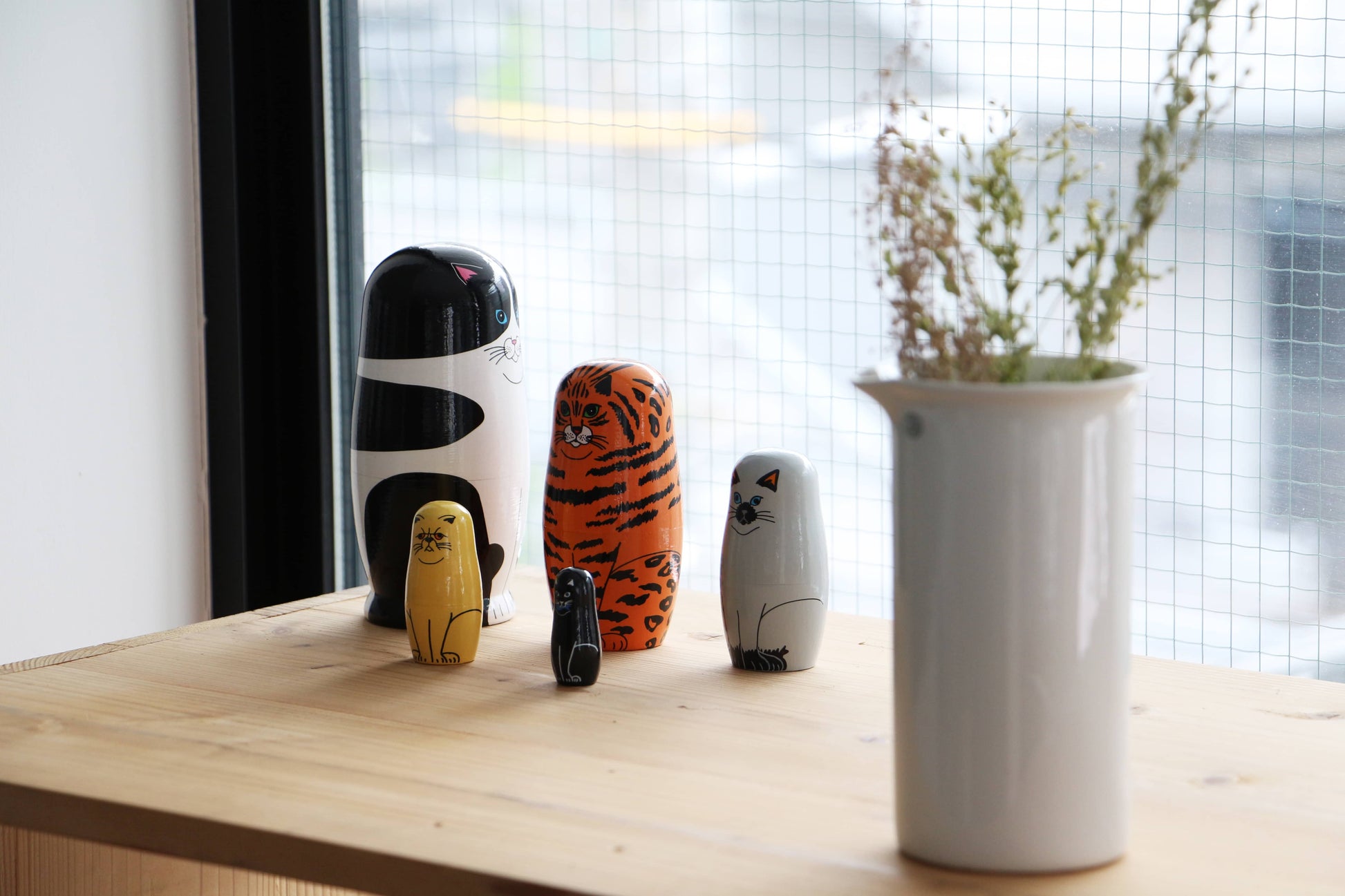 The DETAIL Feline 5 Nesting Dolls by DETAIL, featuring animal-shaped designs like a tiger and cat, are displayed on a wooden surface near a window with a white vase and dried flowers against an urban backdrop—perfect for cat lovers.