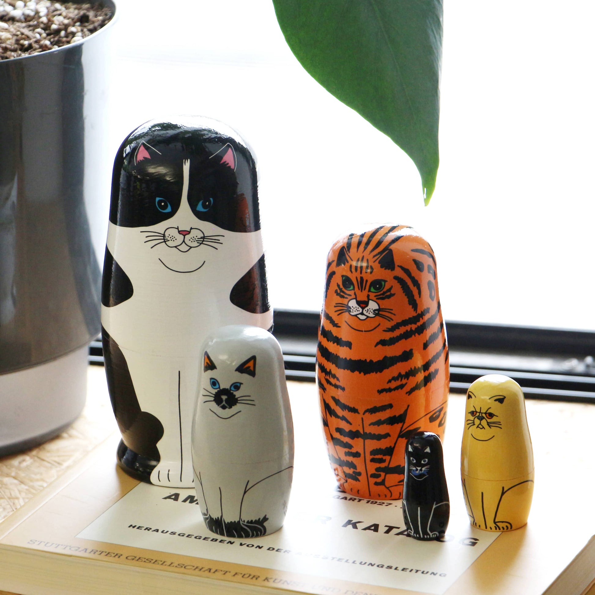 Four DETAIL Feline 5 Nesting Dolls, each with a unique cat design and color, stand on a book near a potted plant. These charming wooden dolls by DETAIL are perfect for cat lovers, accompanied by a green leaf hanging above.