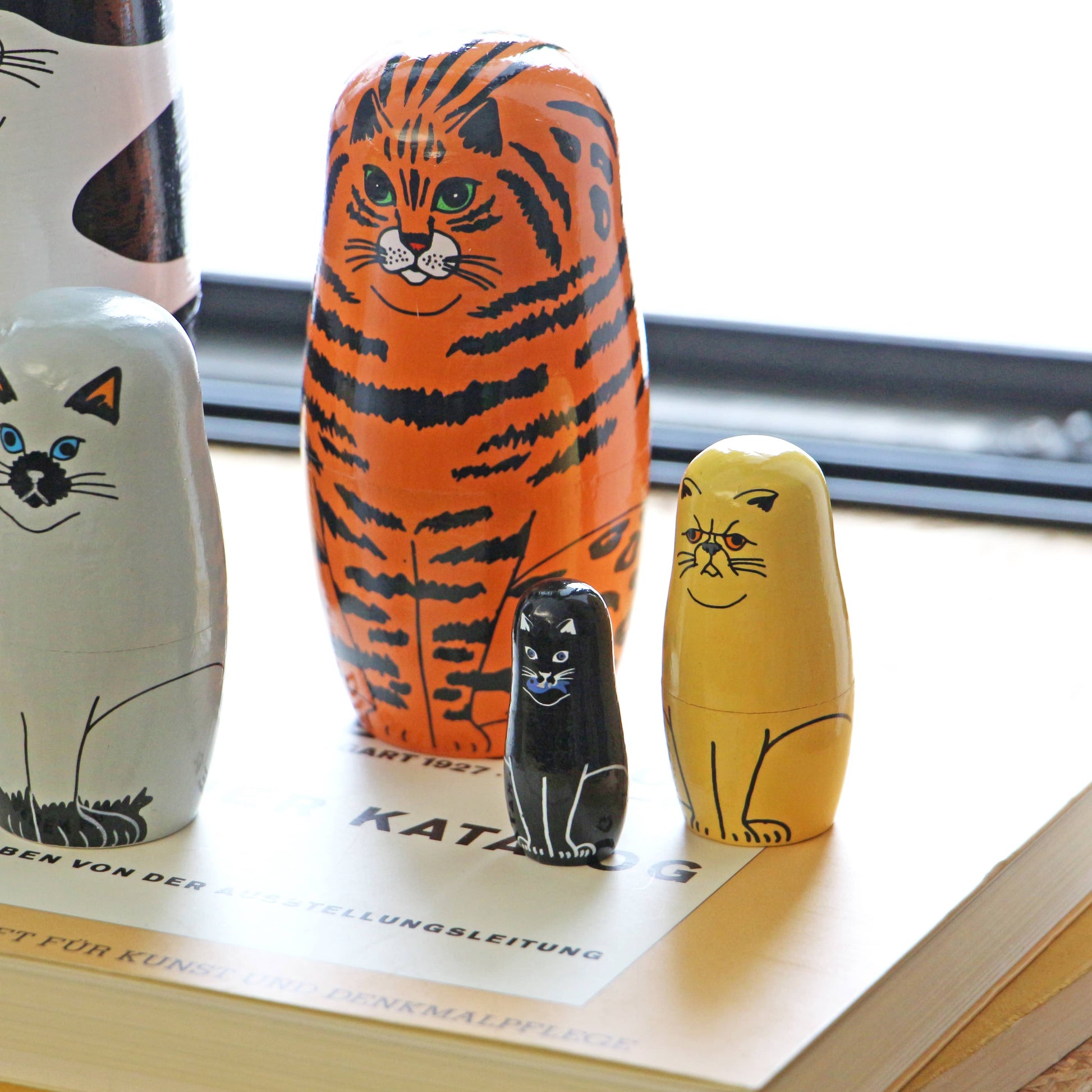 The DETAIL Feline 5 Nesting Dolls by DETAIL, featuring five painted wooden cats in different sizes and colors, are arranged atop a closed book near a window—a delightful display for cat lovers and fans of nesting dolls.