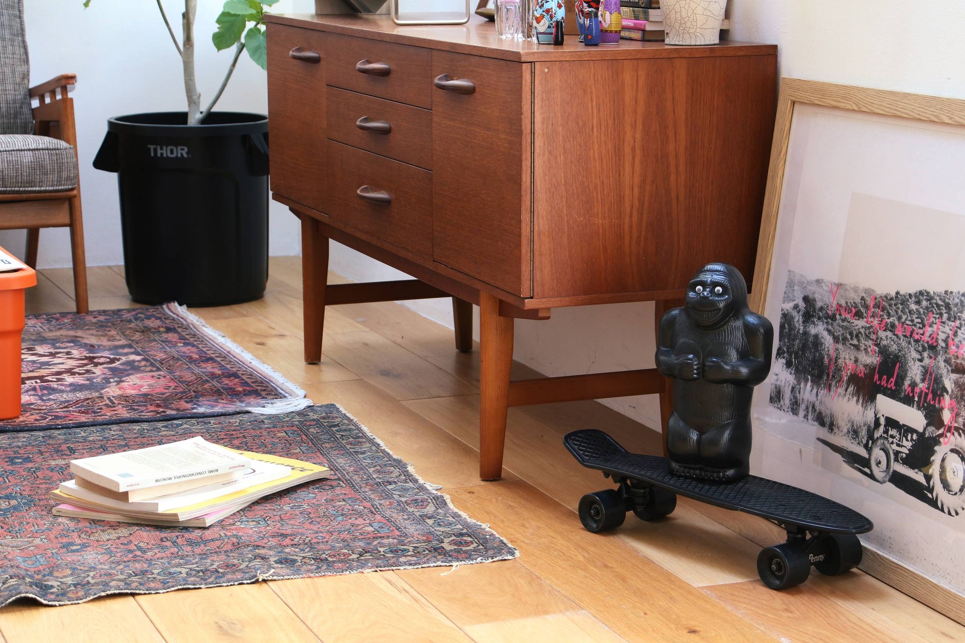 A wooden dresser sits on a hardwood floor near a framed picture, DETAIL Gorilla Bank Small by DETAIL, a skateboard, stacked books, and patterned rugs in a cozy, eclectic room.