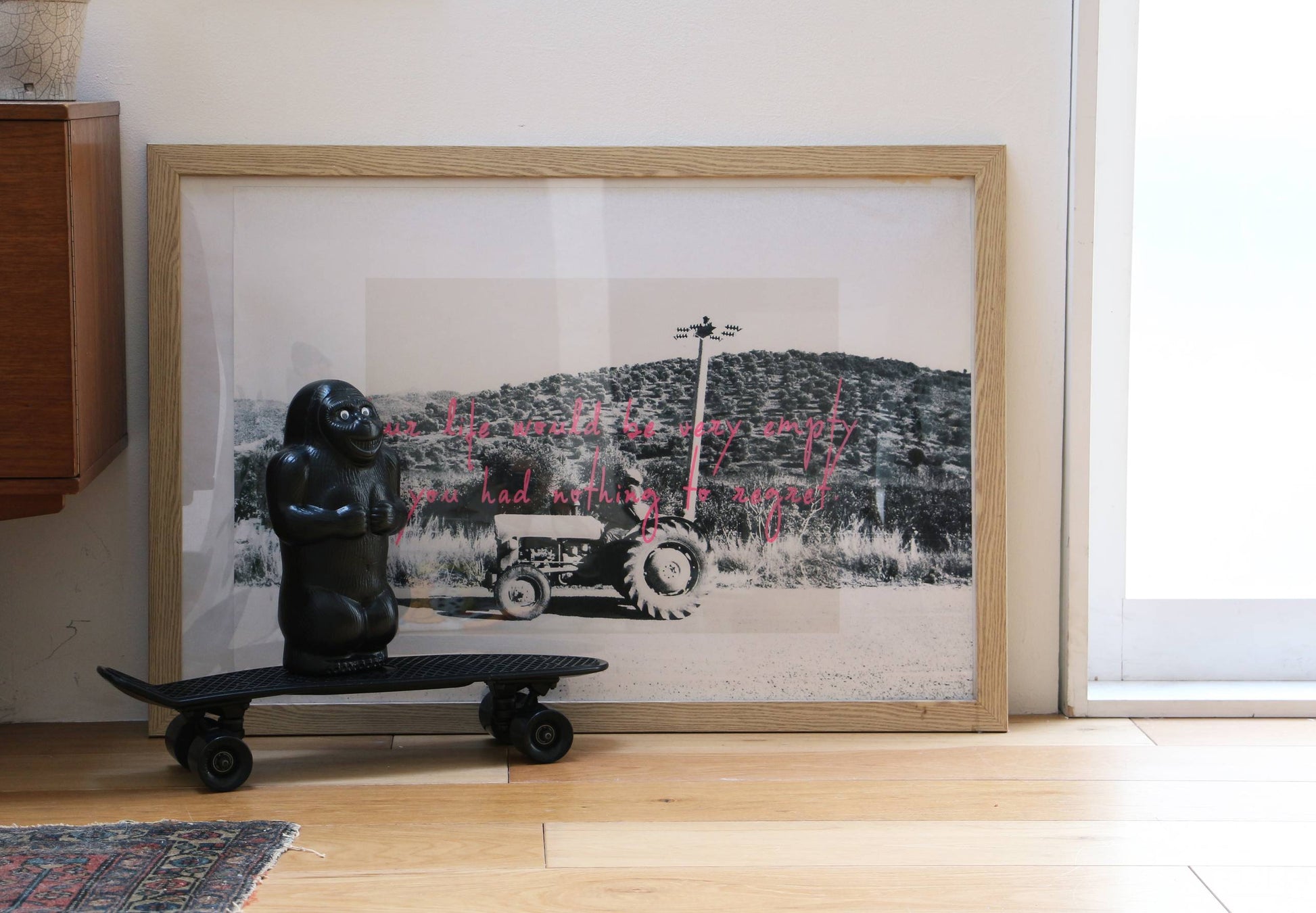 A framed black-and-white tractor photo with pink text stands on a wooden floor near a window. In front, the DETAIL Gorilla Bank Small by DETAIL rides a mini skateboard, adding a playful touch to the decor.