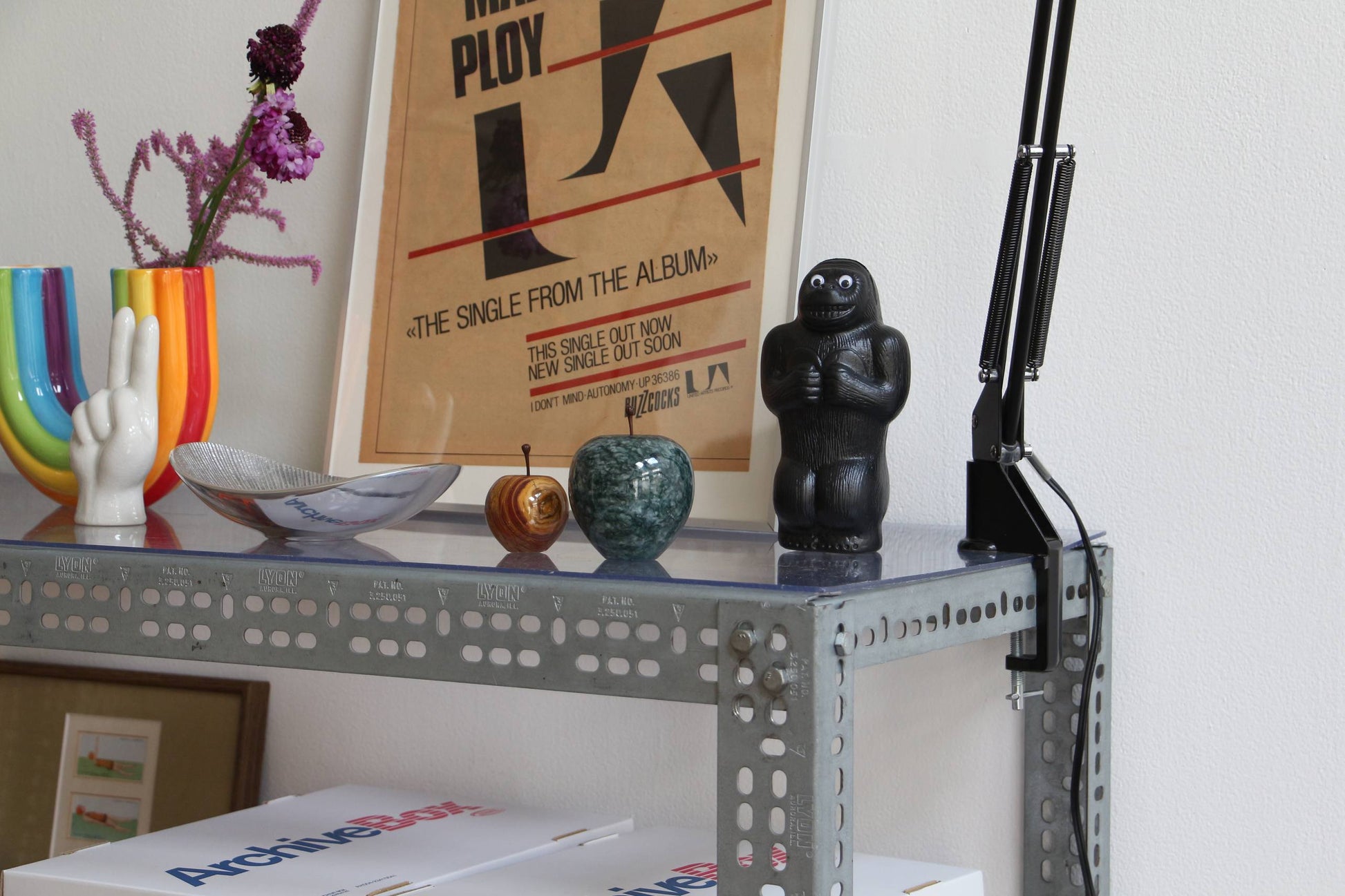 A metal shelf displays colorful decor, including a rainbow vase shaped like a peace-sign hand, the DETAIL Gorilla Bank Small by DETAIL, a lamp, and framed artwork leaning against a white wall.