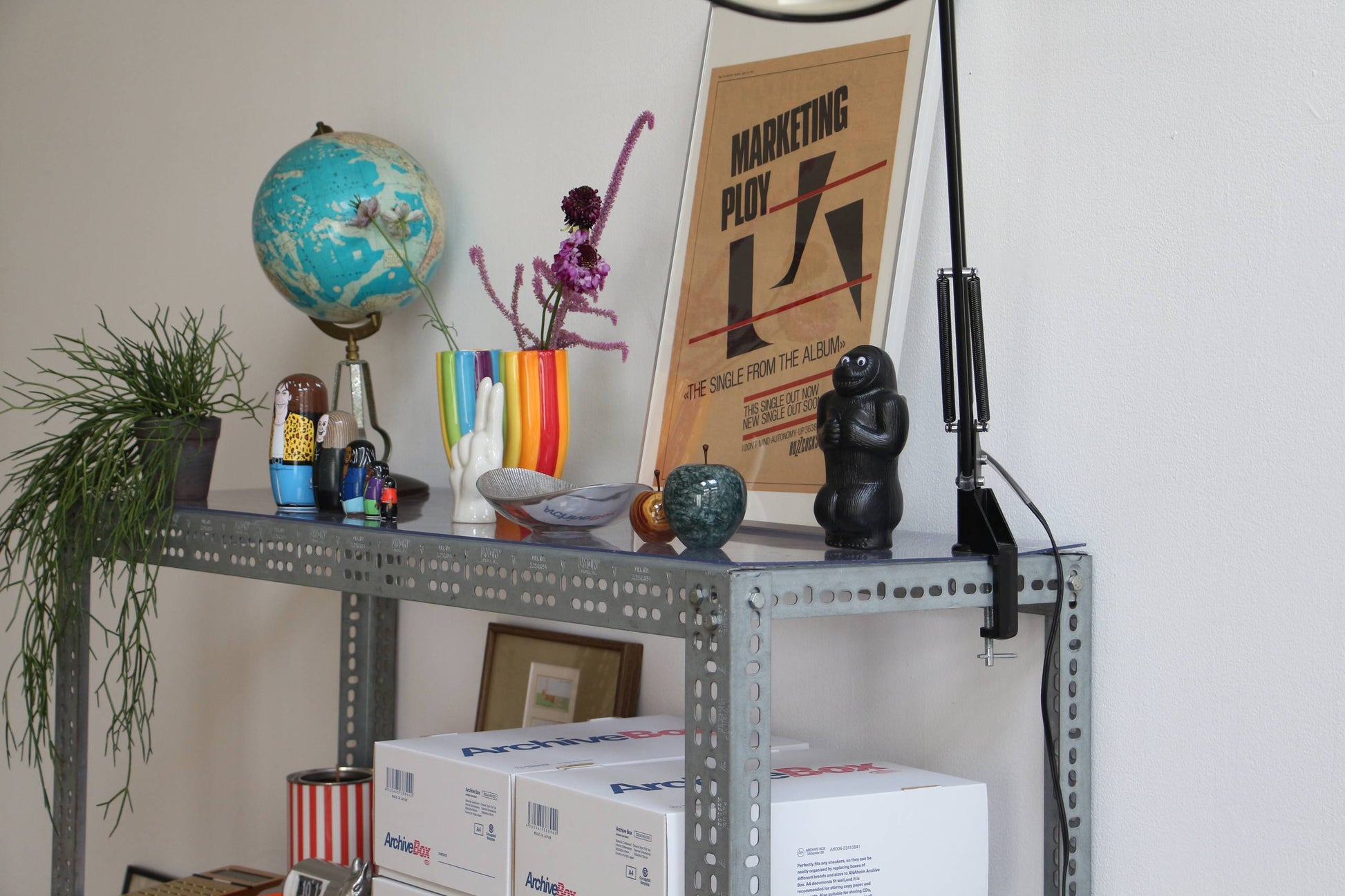 A metal shelf holds a globe, potted plants, colorful vases, the DETAIL Gorilla Bank Small by DETAIL as decor, boxes, a lamp, and a framed MARKETING PLOY poster against a white wall.