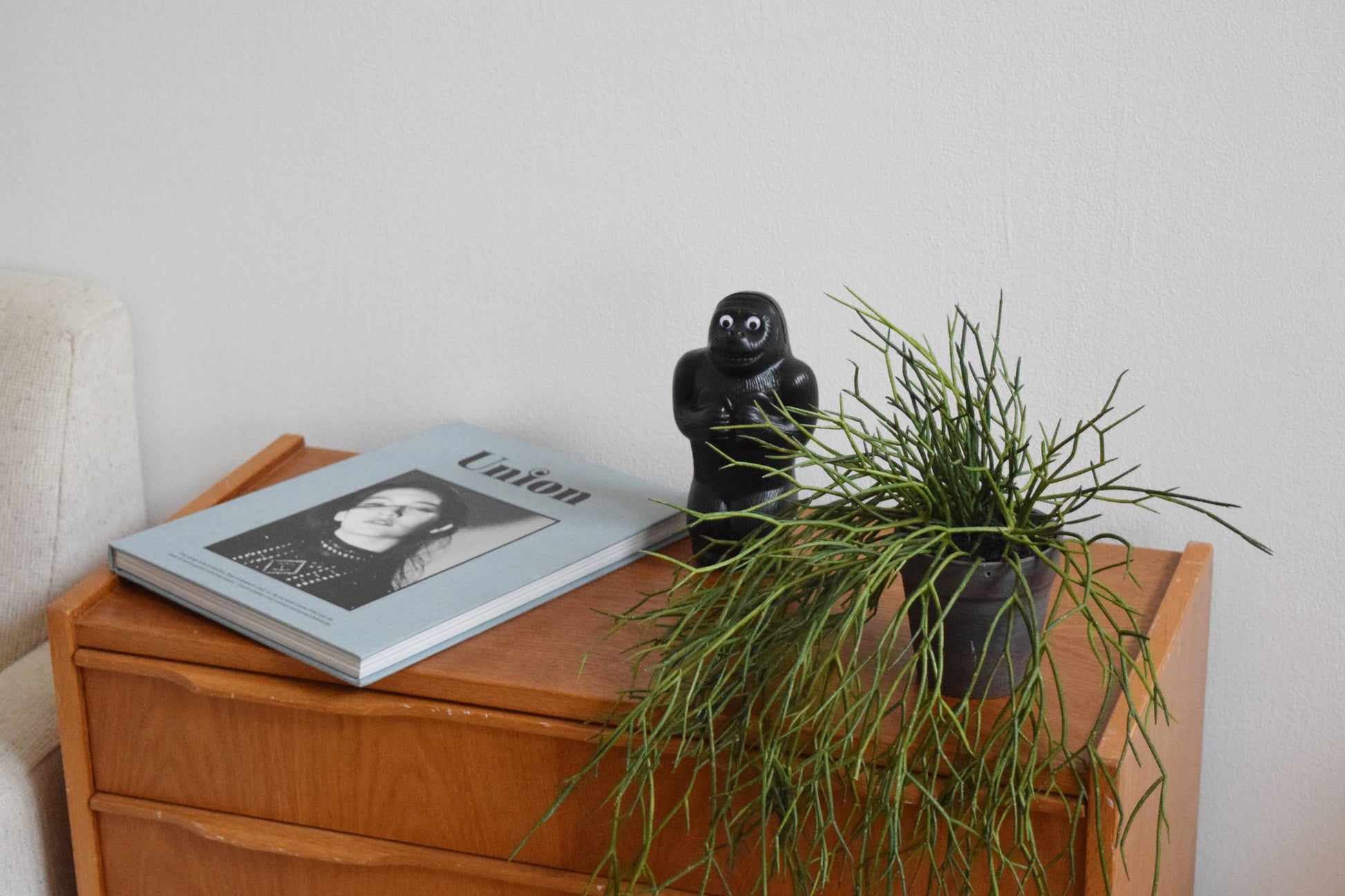 A small wooden side table holds a magazine with a black-and-white portrait on the cover, a DETAIL Gorilla Bank Small by DETAIL, and a potted green plant, set against a plain white wall.