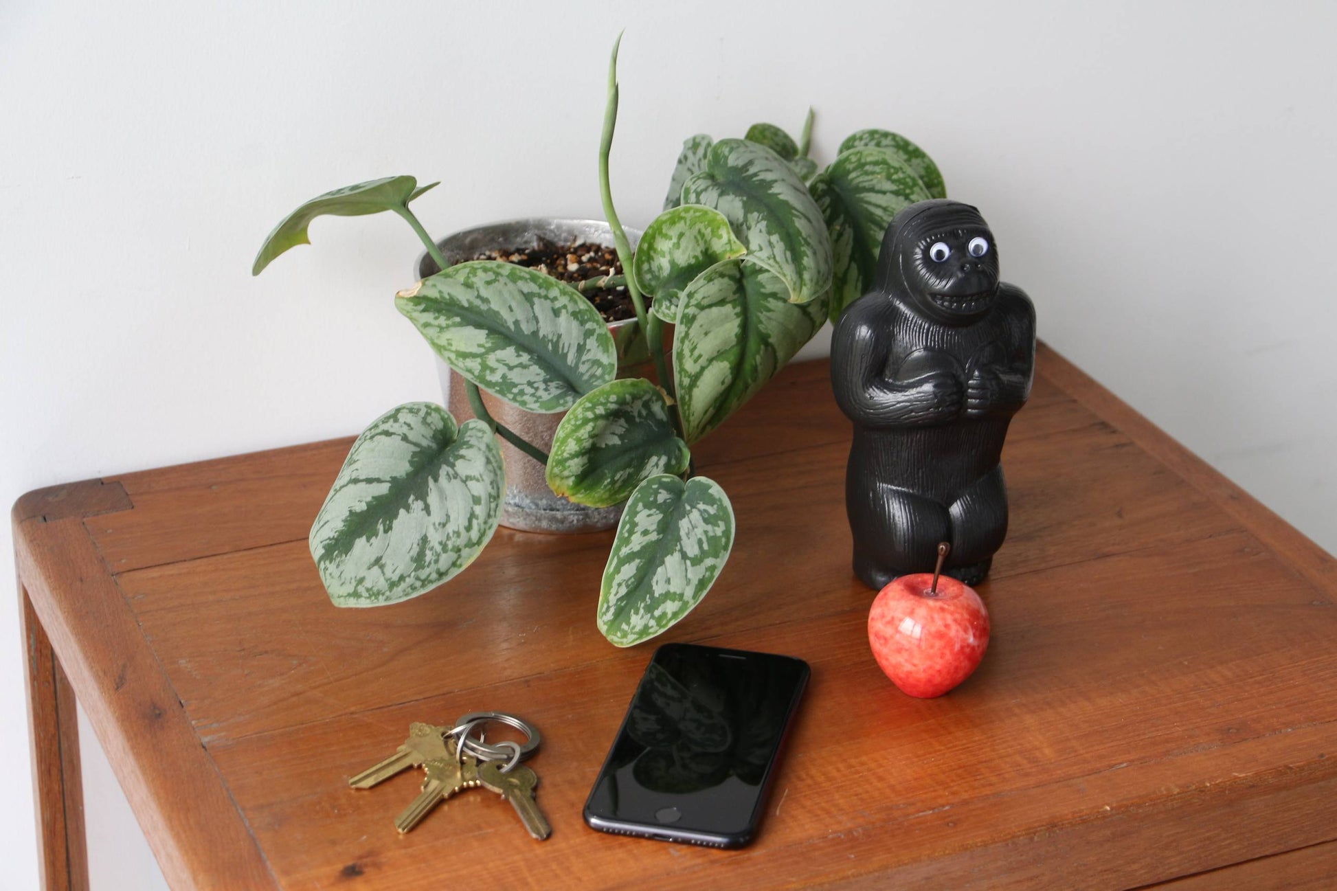 A wooden table with a potted plant, DETAILs Gorilla Bank Small by DETAIL beside a red apple, plus a set of keys and a smartphone on top.