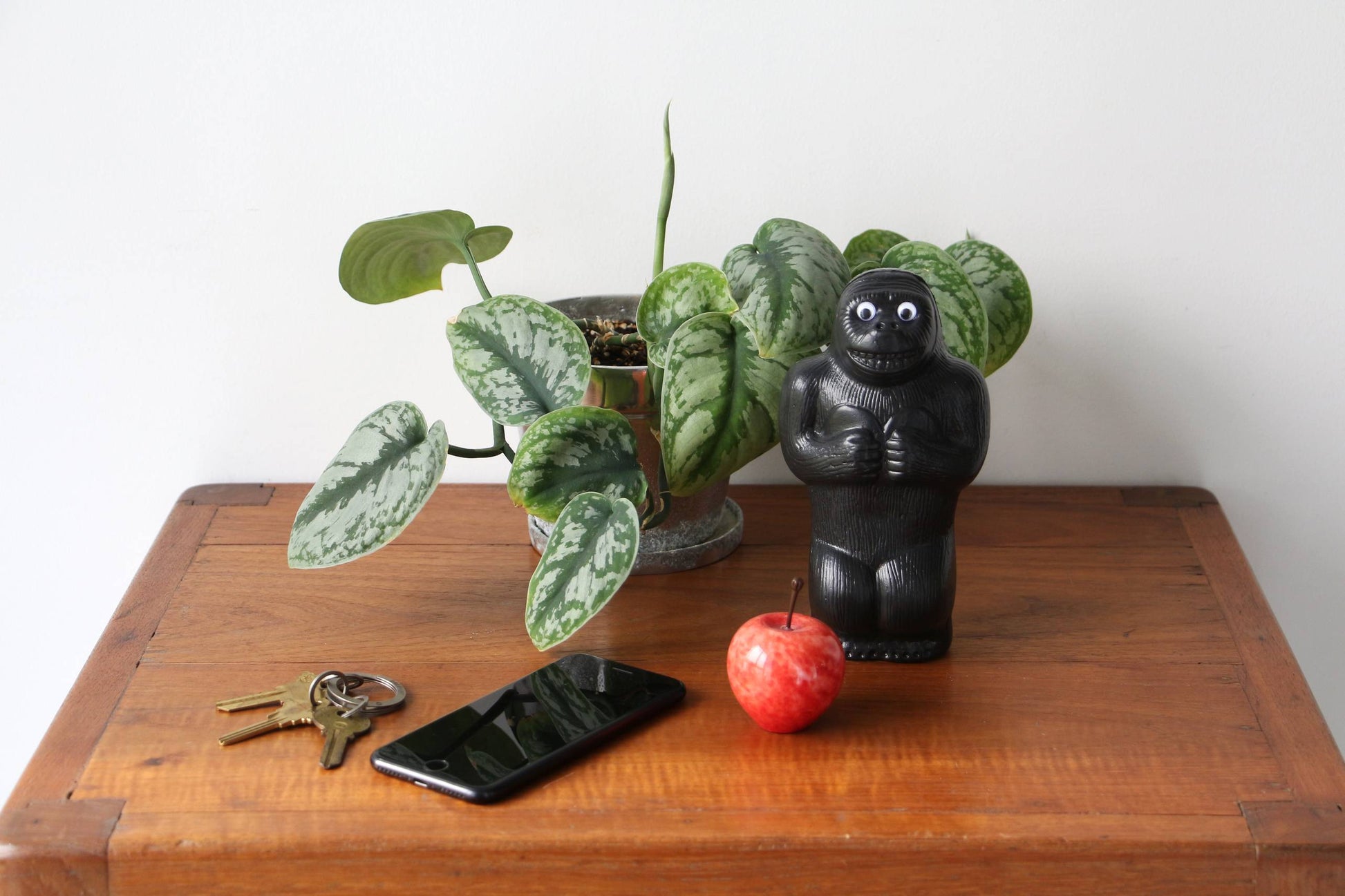 A wooden table with a potted plant, the DETAIL Gorilla Bank Small by DETAIL, a red apple, a smartphone, and a set of keys on top. The background features a plain white wall.