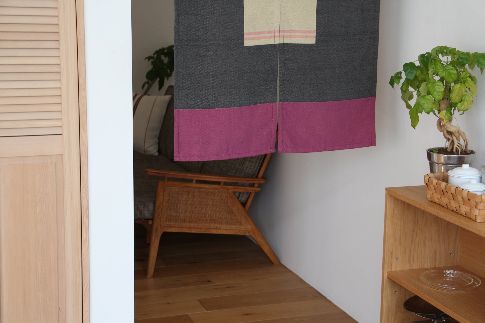 A cozy room features a wooden chair, potted plant, shelves, and a DETAIL Noren Curtain by DETAIL in gray and magenta. The curtain hangs partly open, softly filtering light into the inviting seating area behind it.