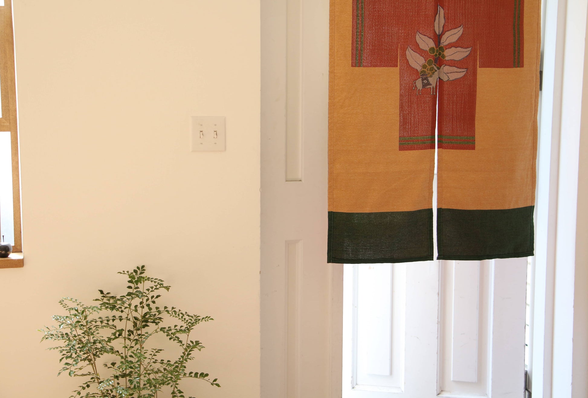 A small leafy green plant sits by a white wall and window. Nearby, a doorway features the DETAIL Noren Curtain from DETAIL, decorated with a plant-inspired yellow and green floral design.