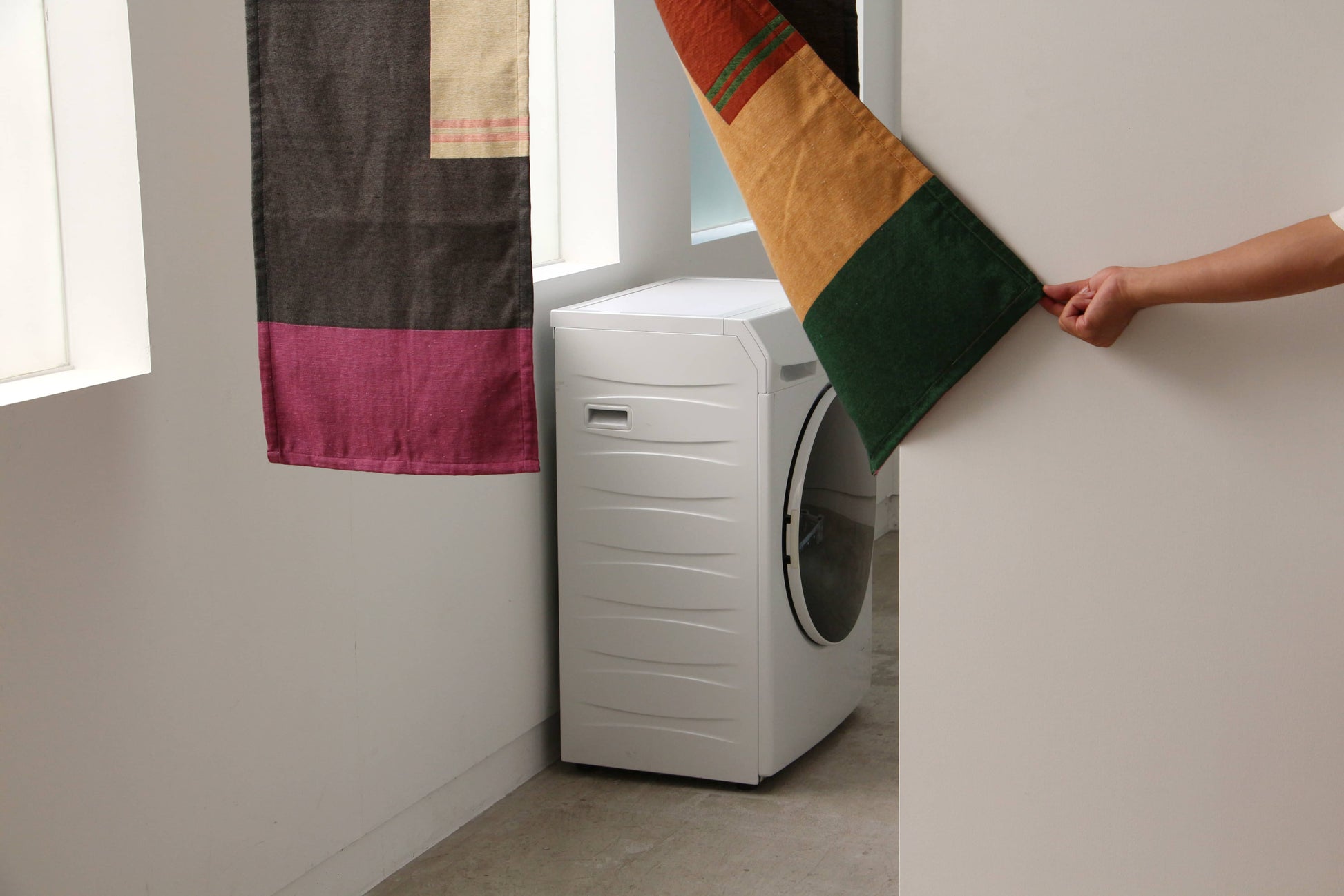 A person lifts a DETAIL Noren Curtain by DETAIL, decorated with kimono patterns, to reveal a white front-loading washing machine in a bright, narrow room with white walls and large windows.