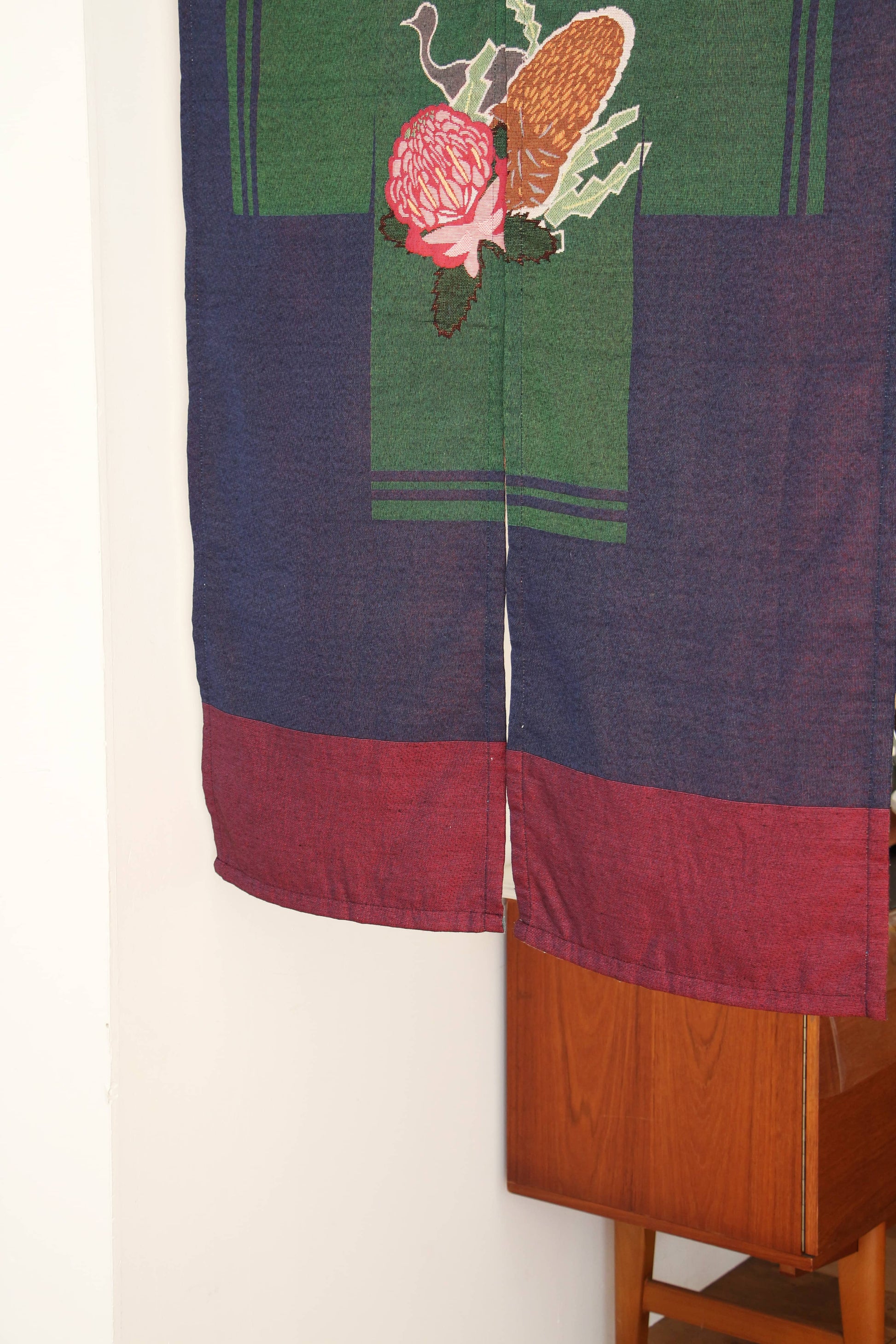 The DETAIL Noren Curtain by DETAIL features a red and navy bottom border and a green panel with plant-inspired florals and pinecones, hanging partially open near a wooden cabinet against a white wall.